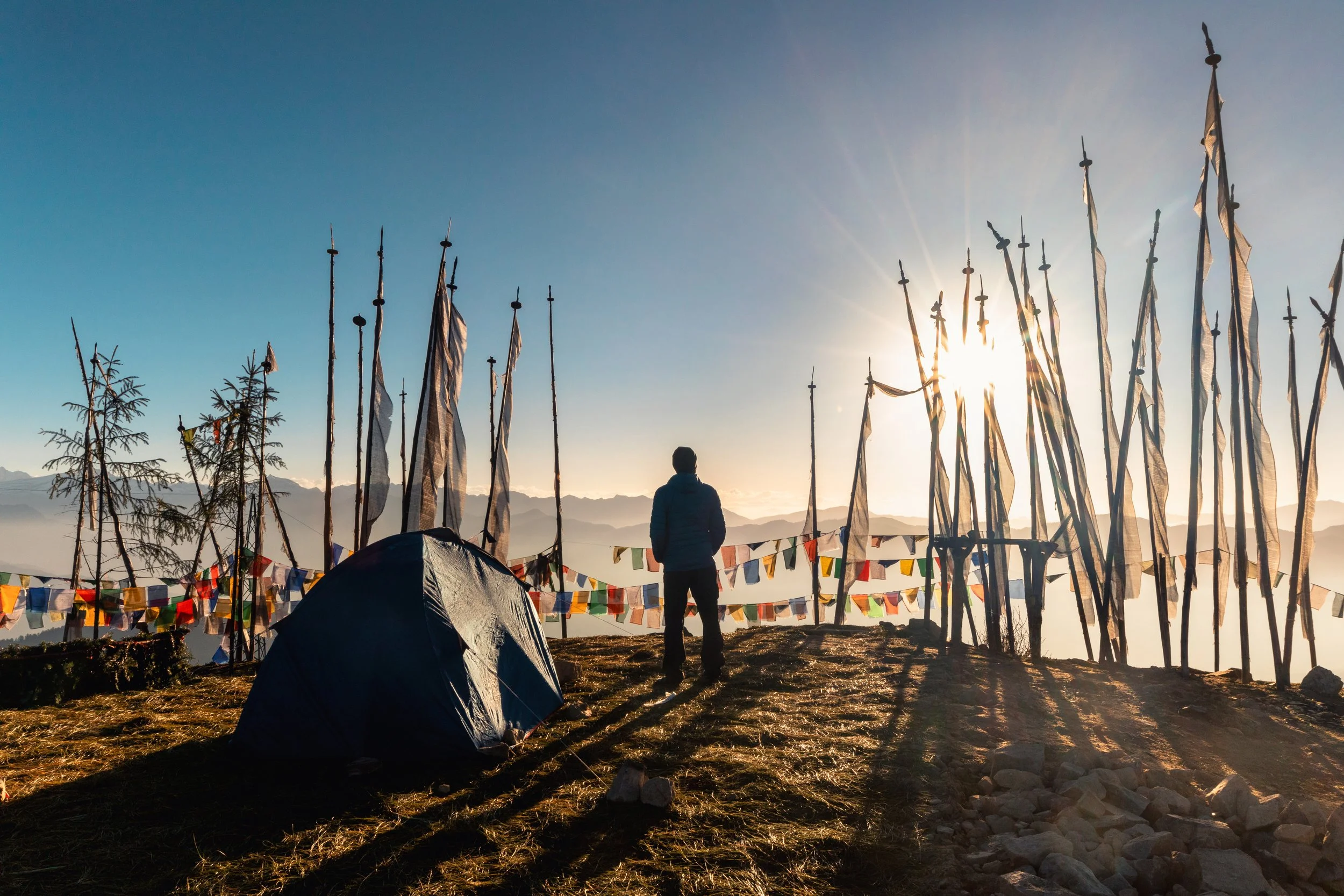 A person stands in a field during sunset, surrounded by prayer flags on poles and a small tent, with mountains in the background.