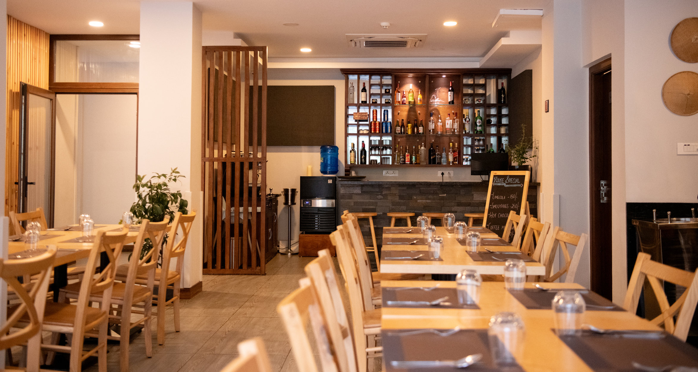 Interior of a cozy restaurant with wooden tables and chairs, a bar with bottles of liquor, a water dispenser, and a potted plant, illuminated with warm lighting.