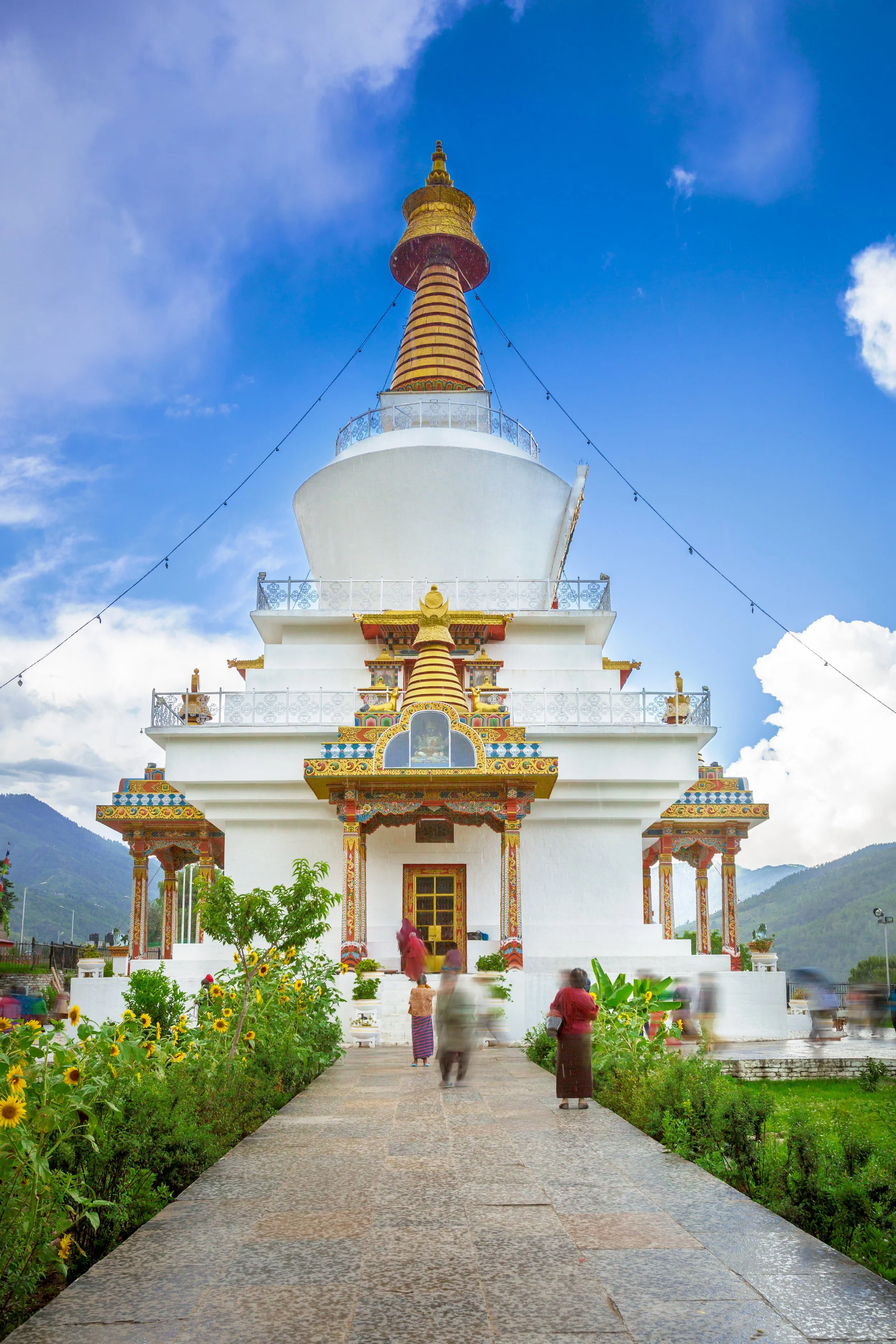 A white Buddhist temple with colorful ornate decorations and golden accents. The temple has a tiered roof and a tall central stupa with a golden spire. There are people walking towards the temple entrance, surrounded by a garden with yellow sunflowers and green plants, with mountains and a partly cloudy sky in the background.