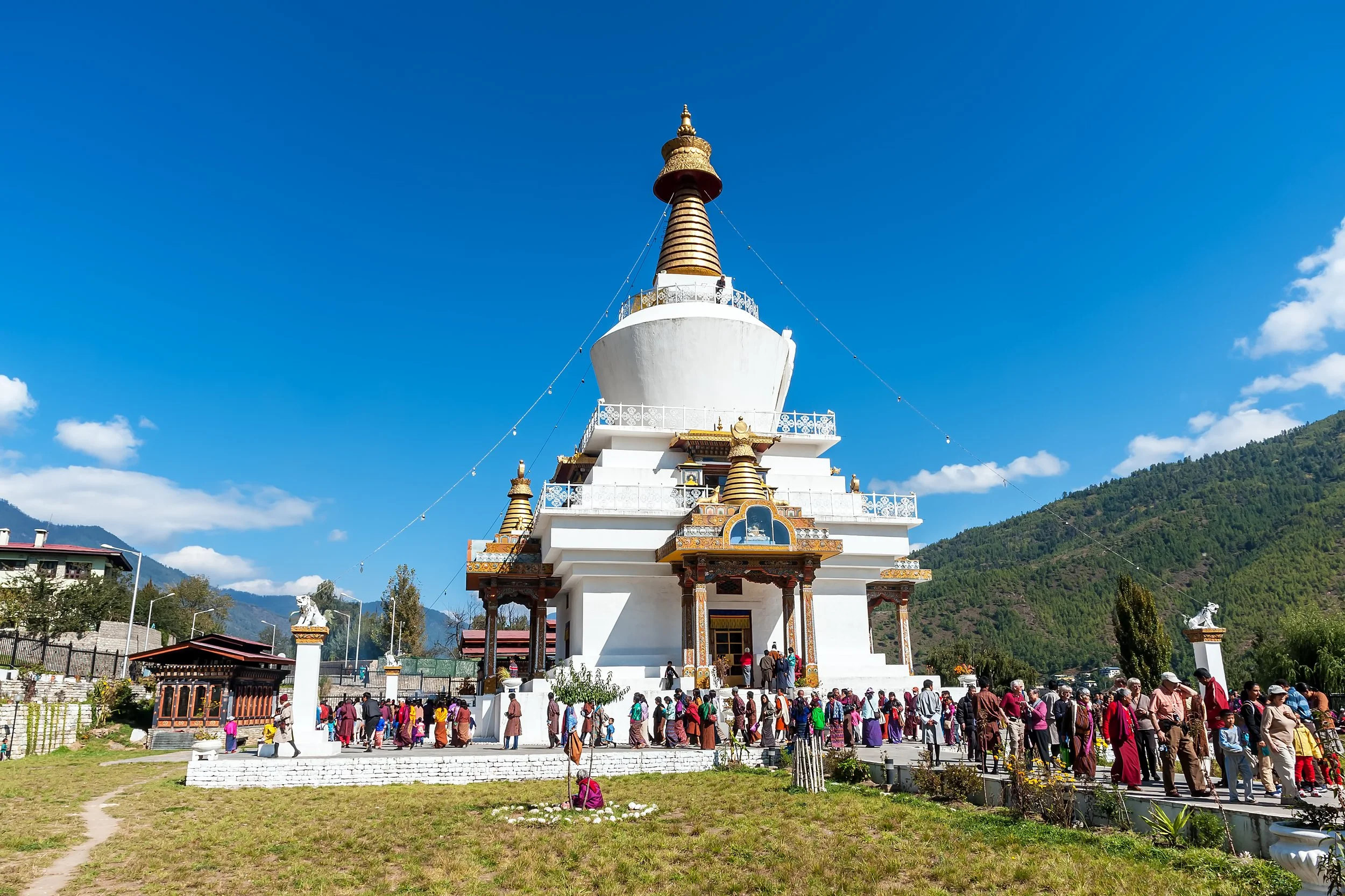 A white Buddhist stupa with golden decorations and multiple smaller golden stupas, set against a backdrop of green hills and a bright blue sky. A large crowd of people is gathered around the base of the stupa.