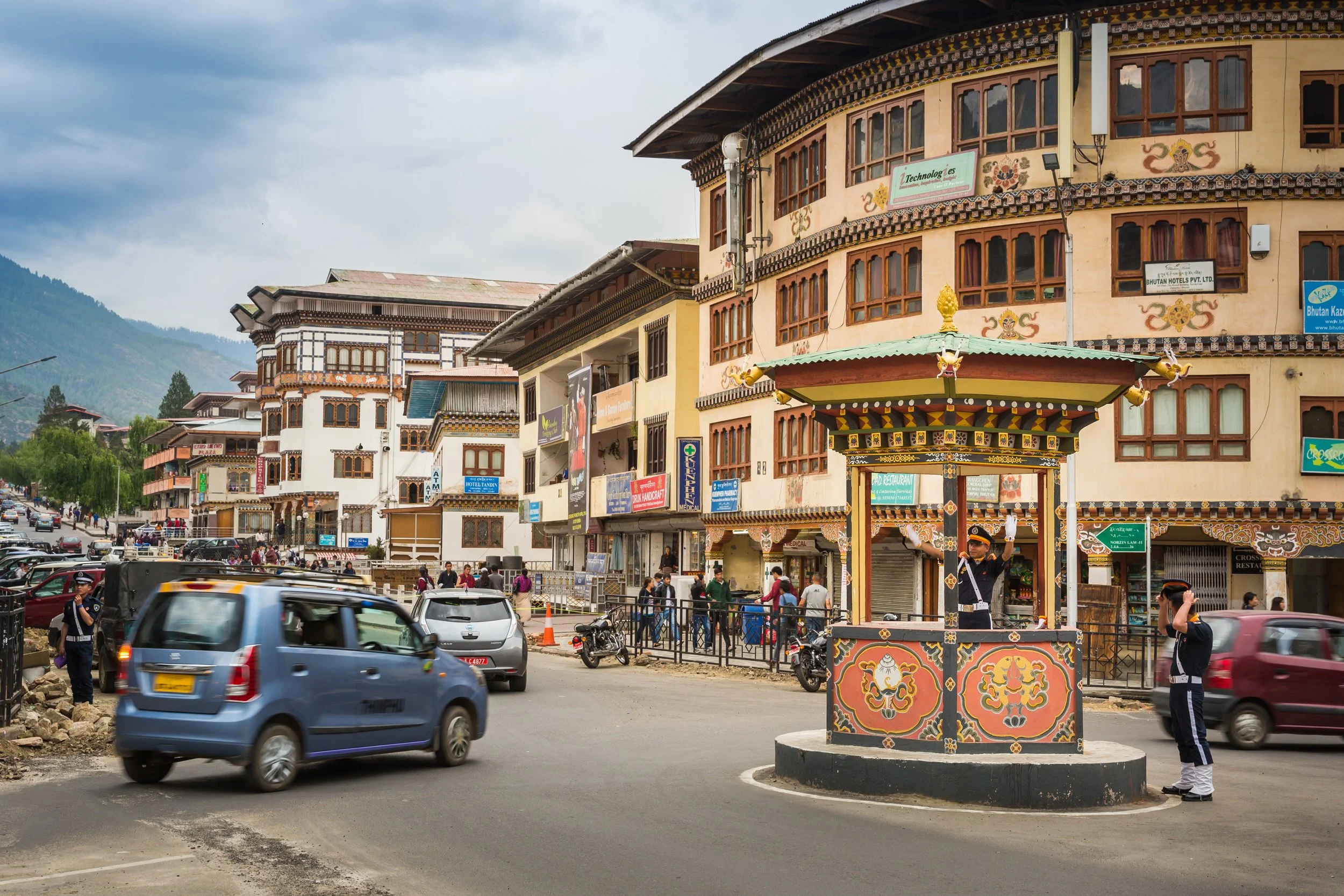 A busy street scene in a town with colorful buildings featuring traditional Bhutanese architecture, and a small decorative structure in the foreground. Cars and people are visible, with mountains in the background.