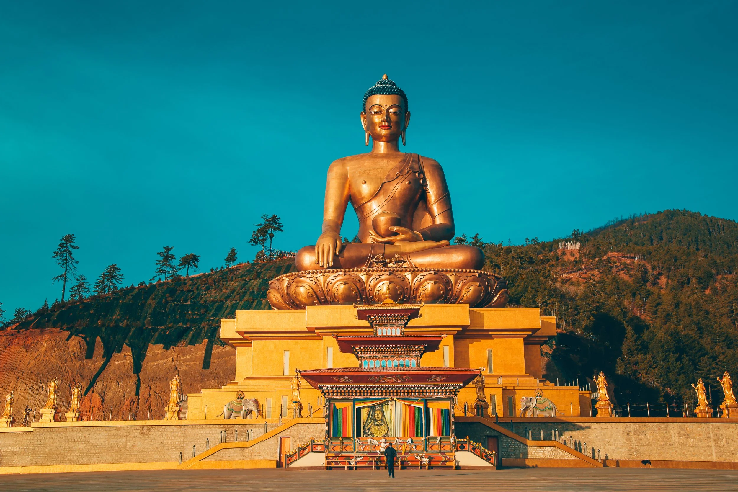 Large golden statue of Buddha seated on a lotus flower pedestal with a mountainous background and trees, under a clear blue sky.