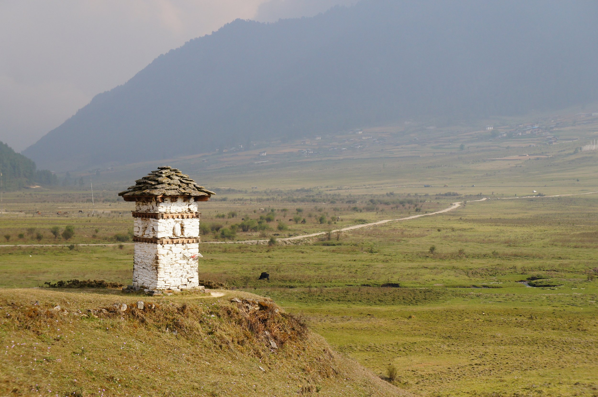 A small stone and brick structure with a shingled roof stands in a vast green field. In the background, rolling hills and mountains are shrouded in mist or haze, with small houses scattered across the landscape.