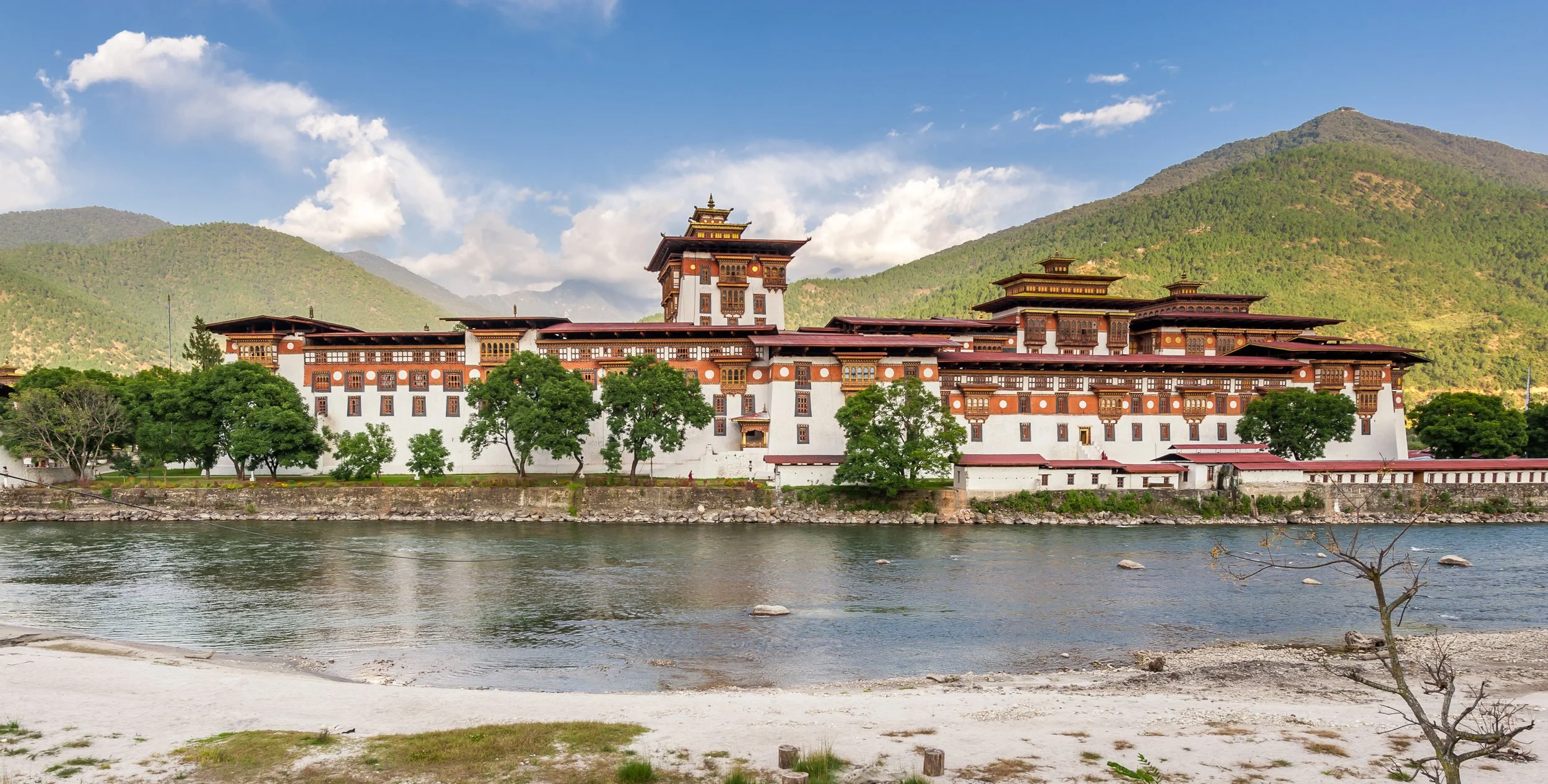 Traditional Bhutanese fortress or dzong by a river with green mountains in the background under a partly cloudy sky.