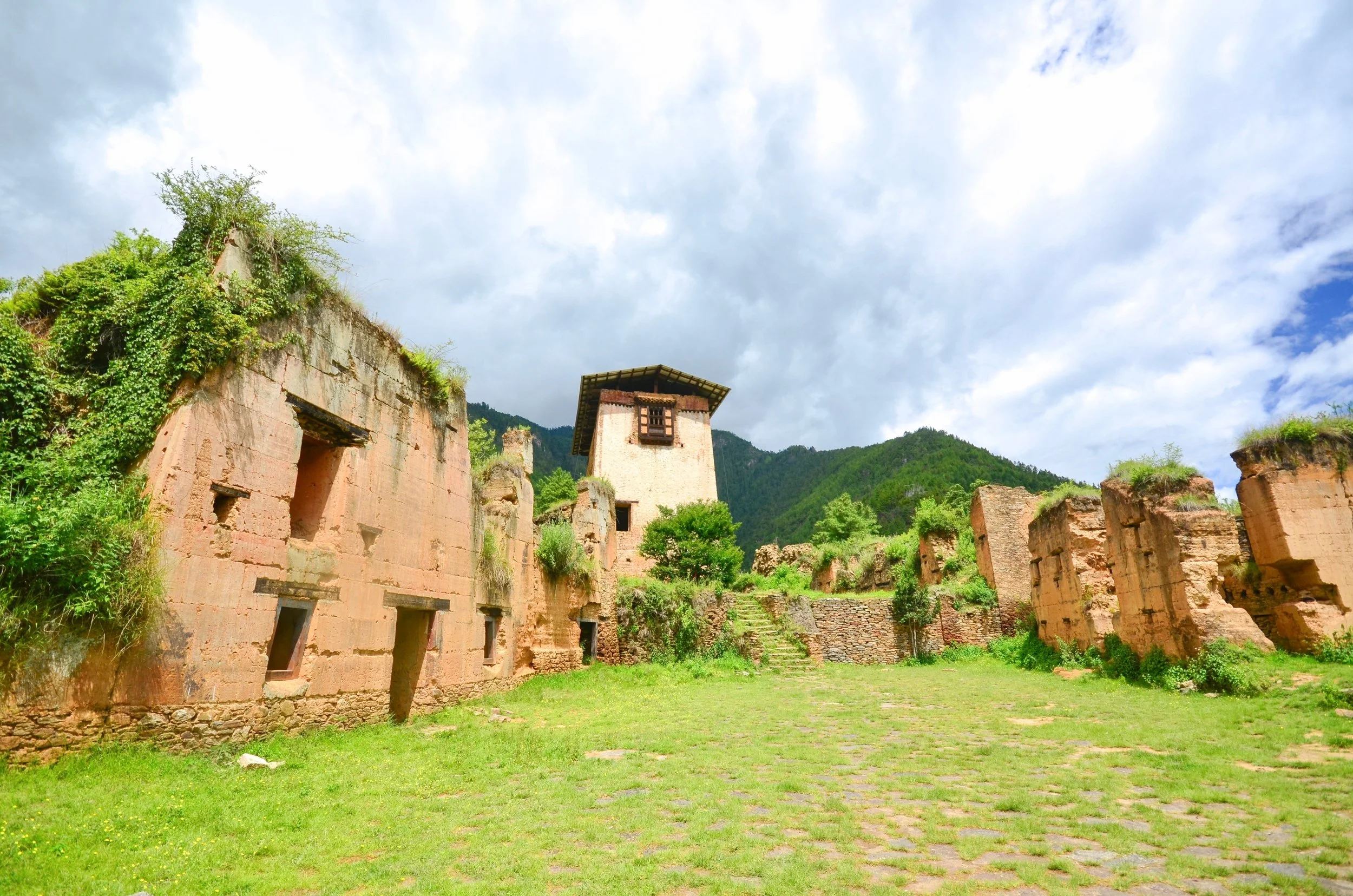 Ancient ruins of stone buildings with greenery on top, set against a backdrop of green mountains and a cloudy sky.