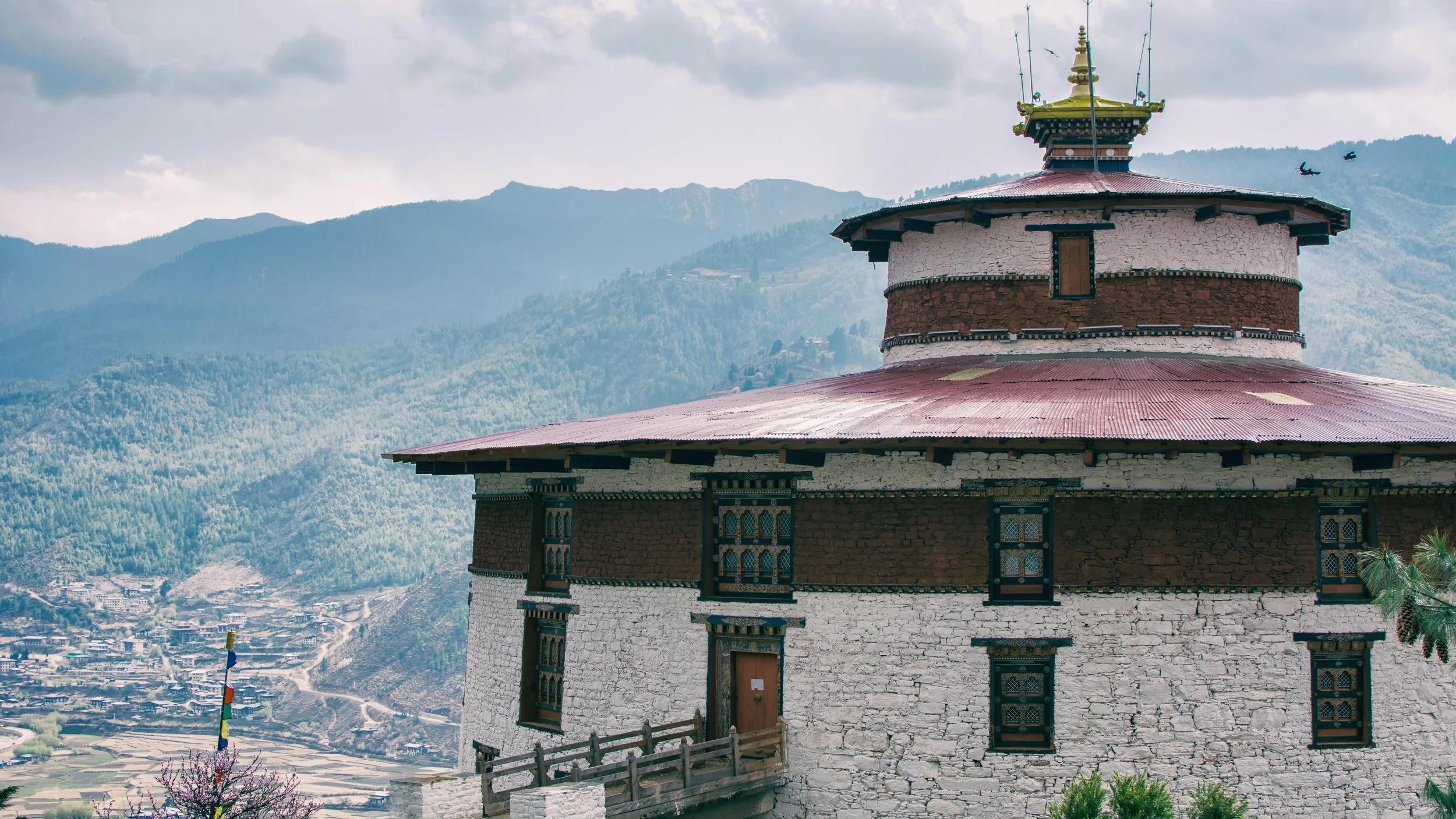 A traditional multi-storied building with white and dark brown walls, and decorative windows, sits on a hillside with mountainous terrain in the background. The building has a red metal roof and ornate details, with a foggy or cloudy sky overhead.