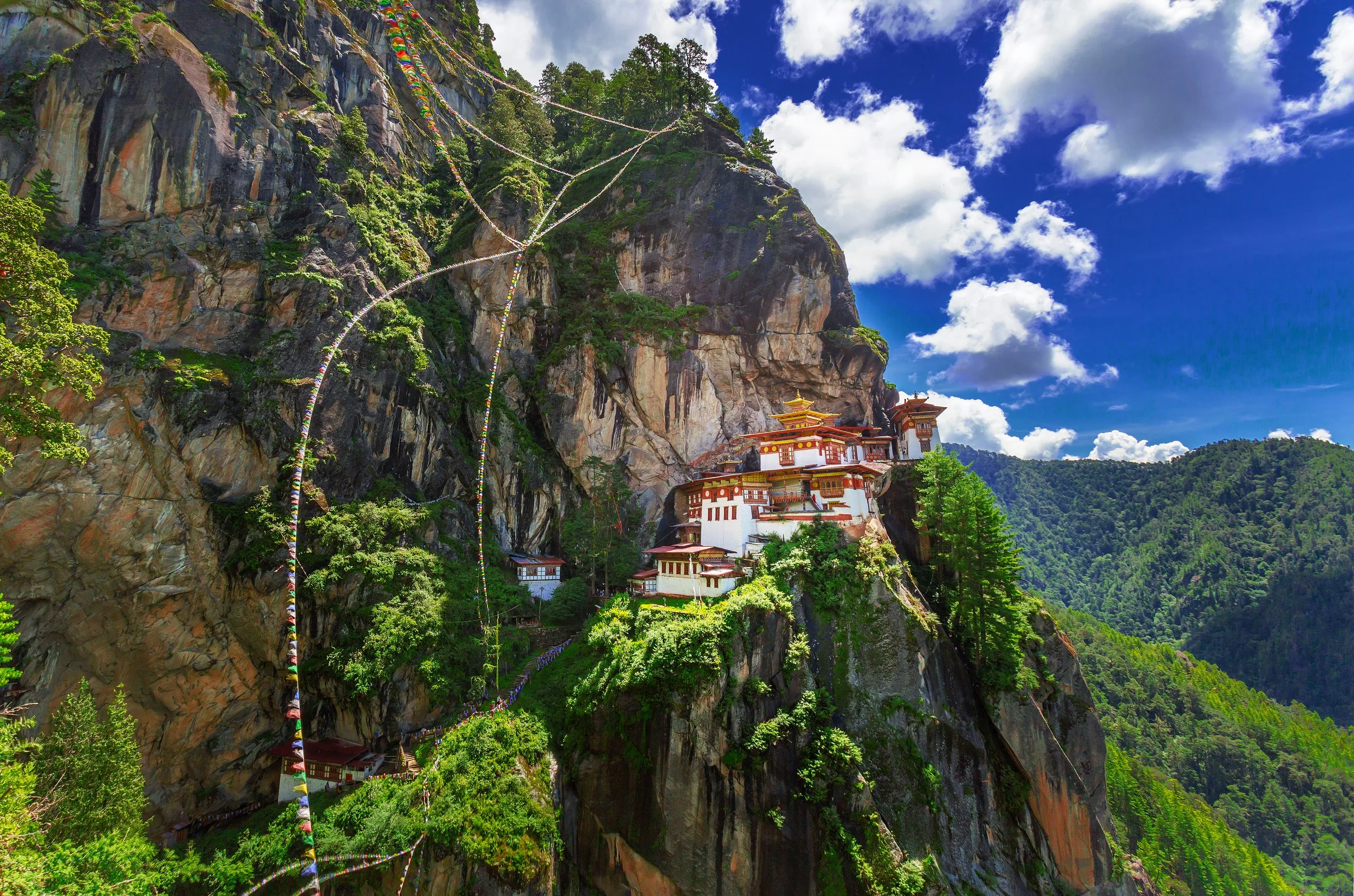 A mountain with a Buddhist temple built into it, colorful prayer flags hanging across the rocks, and a bright blue sky with scattered white clouds.