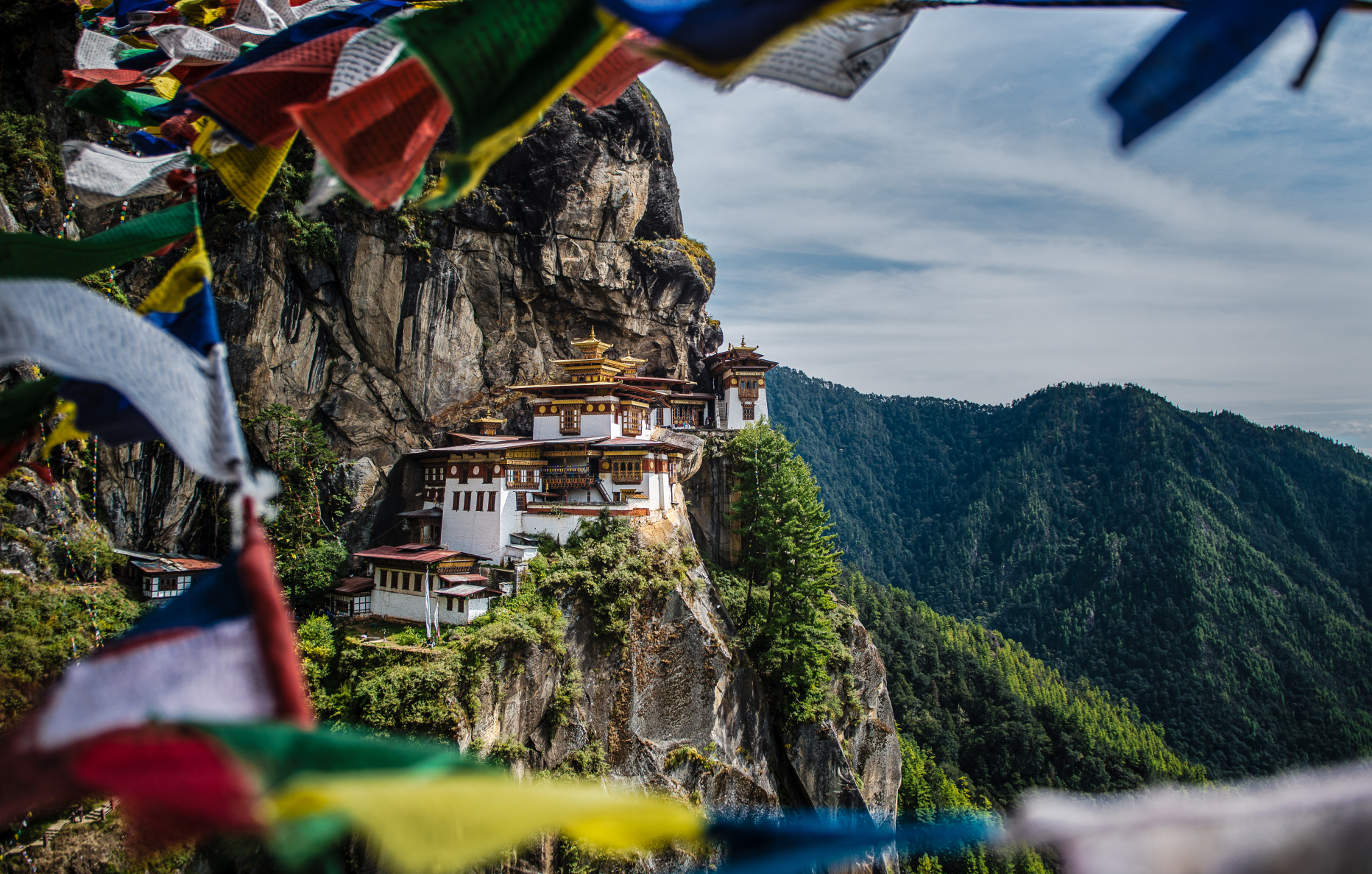 Picture of Tiger's Nest Monastery climbing up a rocky cliffside, with prayer flags in the foreground and forested mountains in the background.