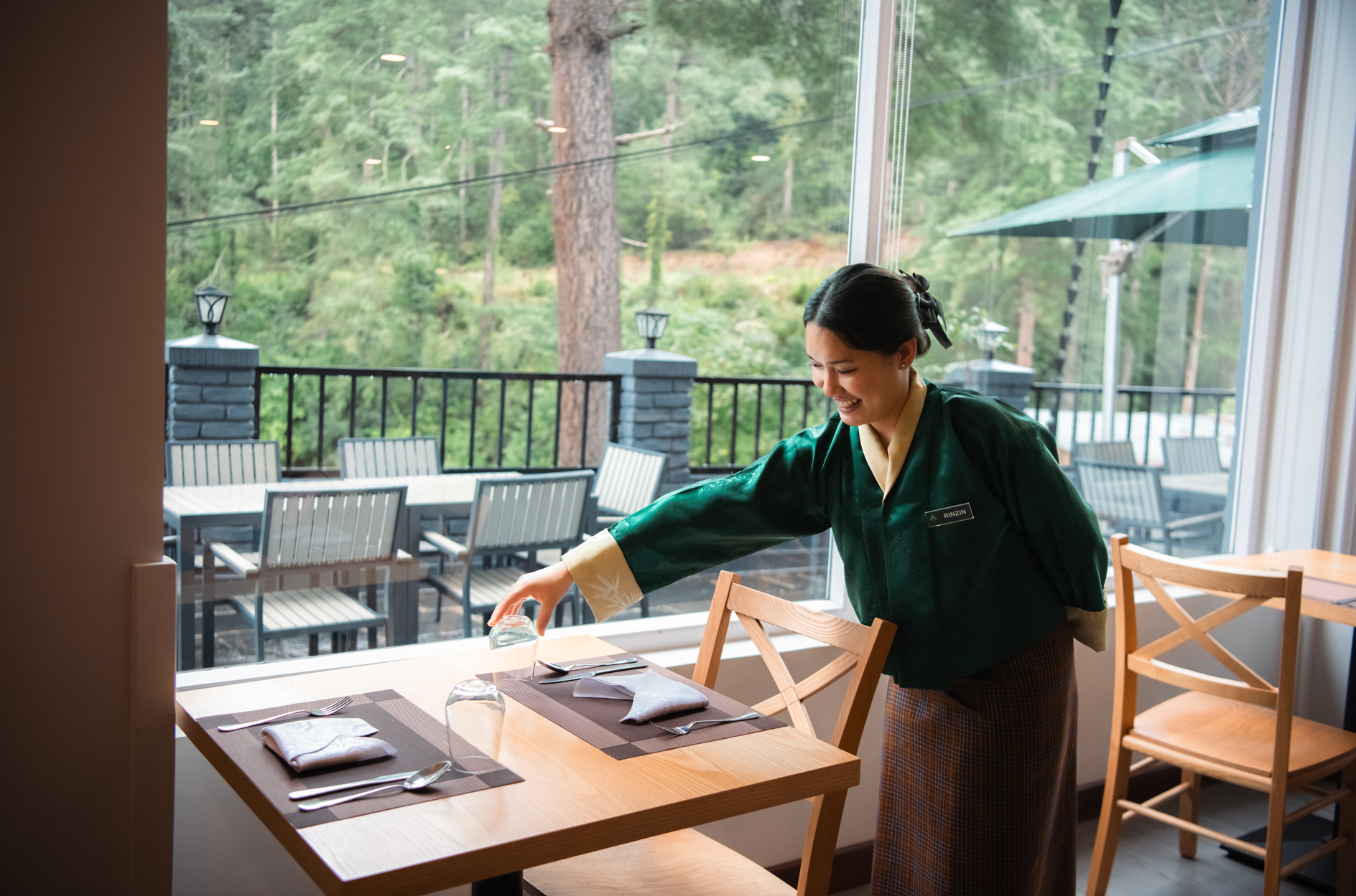 A woman in a green and yellow traditional dress is setting a table in a restaurant with large windows showing a wooded outdoor patio.