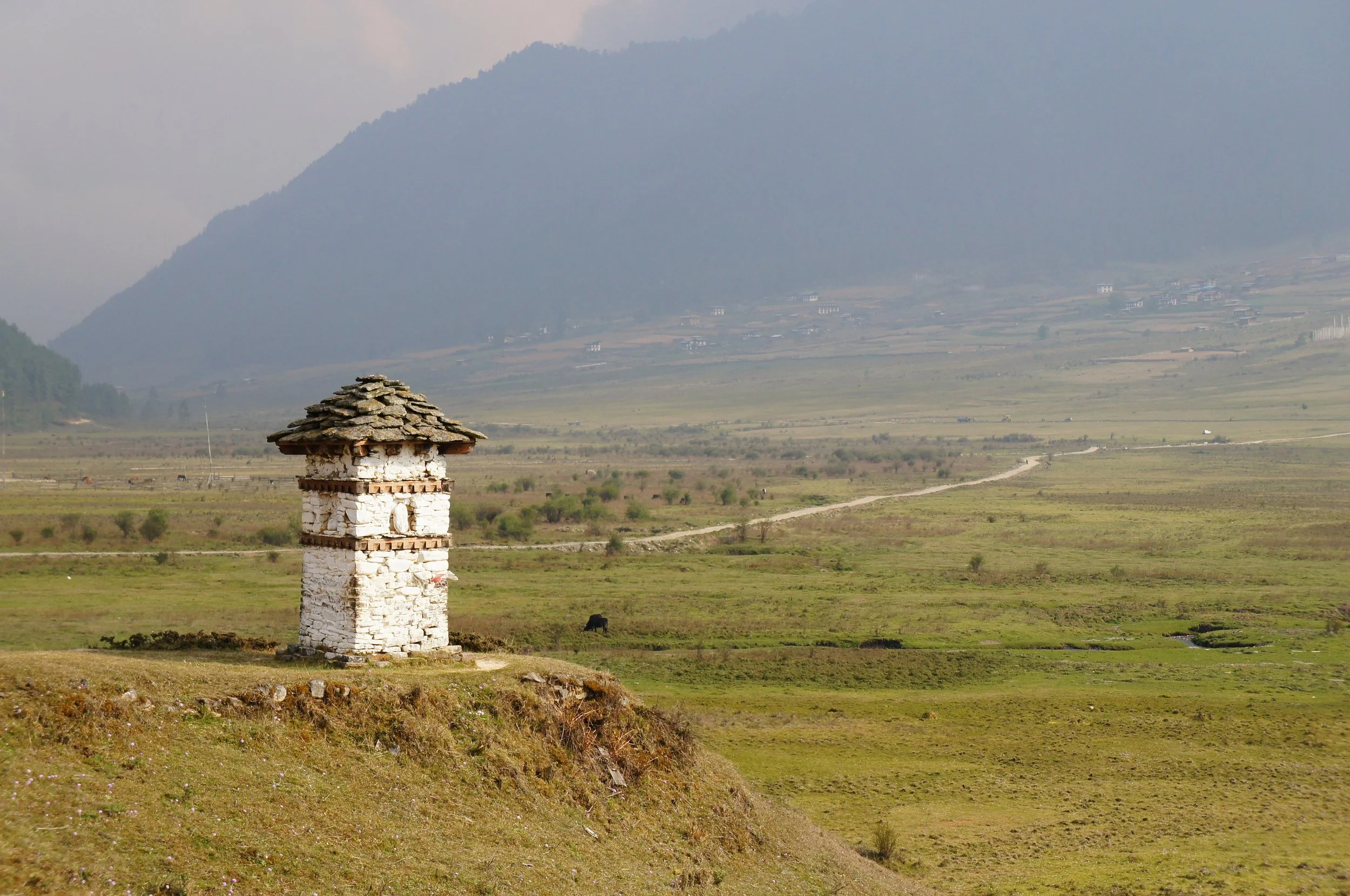 A rural landscape with a white stone tower in the foreground, grassy fields, a dirt path, and distant small houses with mountains and clouds in the background.