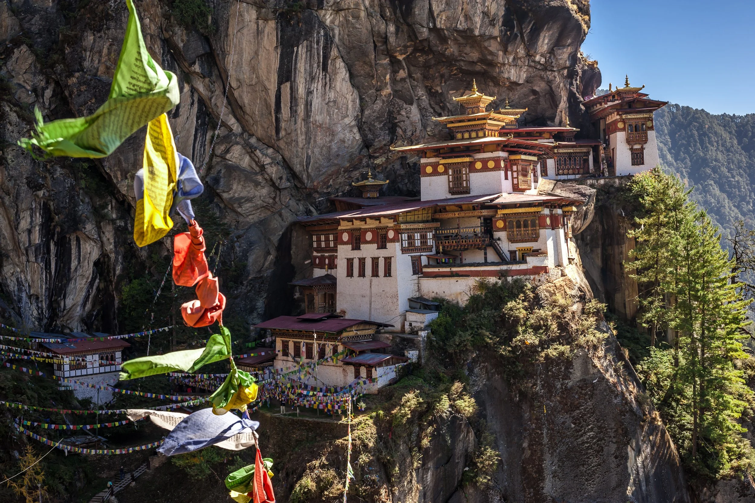 Buddhist monastery built into a rocky mountainside with colorful prayer flags hanging across the scene.