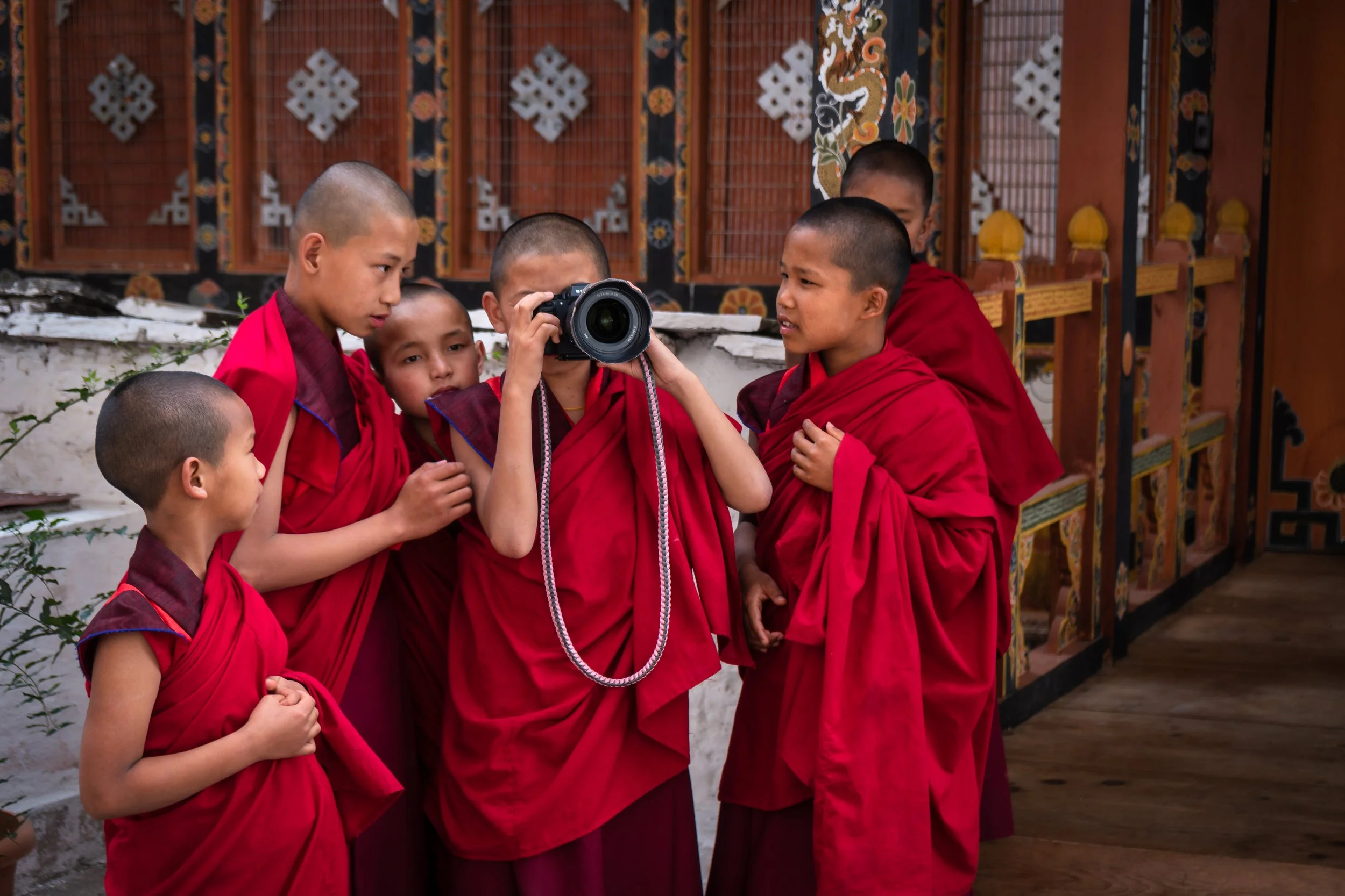 Group of young Buddhist monks in red robes gathered around a camera, looking at its viewfinder, against a decorative background with traditional patterned wood and metal panels.