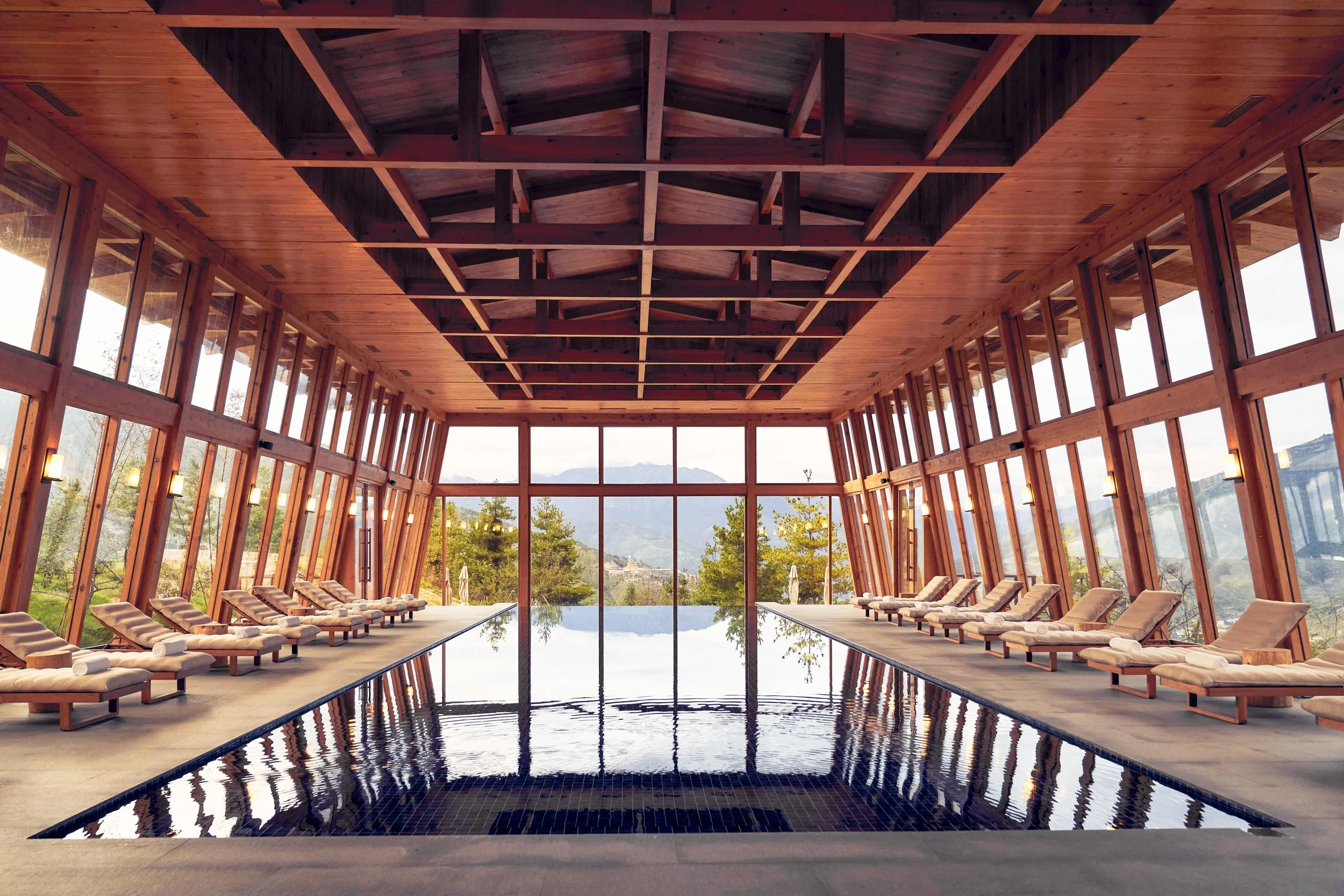Indoor pool area with wooden walls, large windows, lounge chairs along the pool edge, and mountains visible outside.