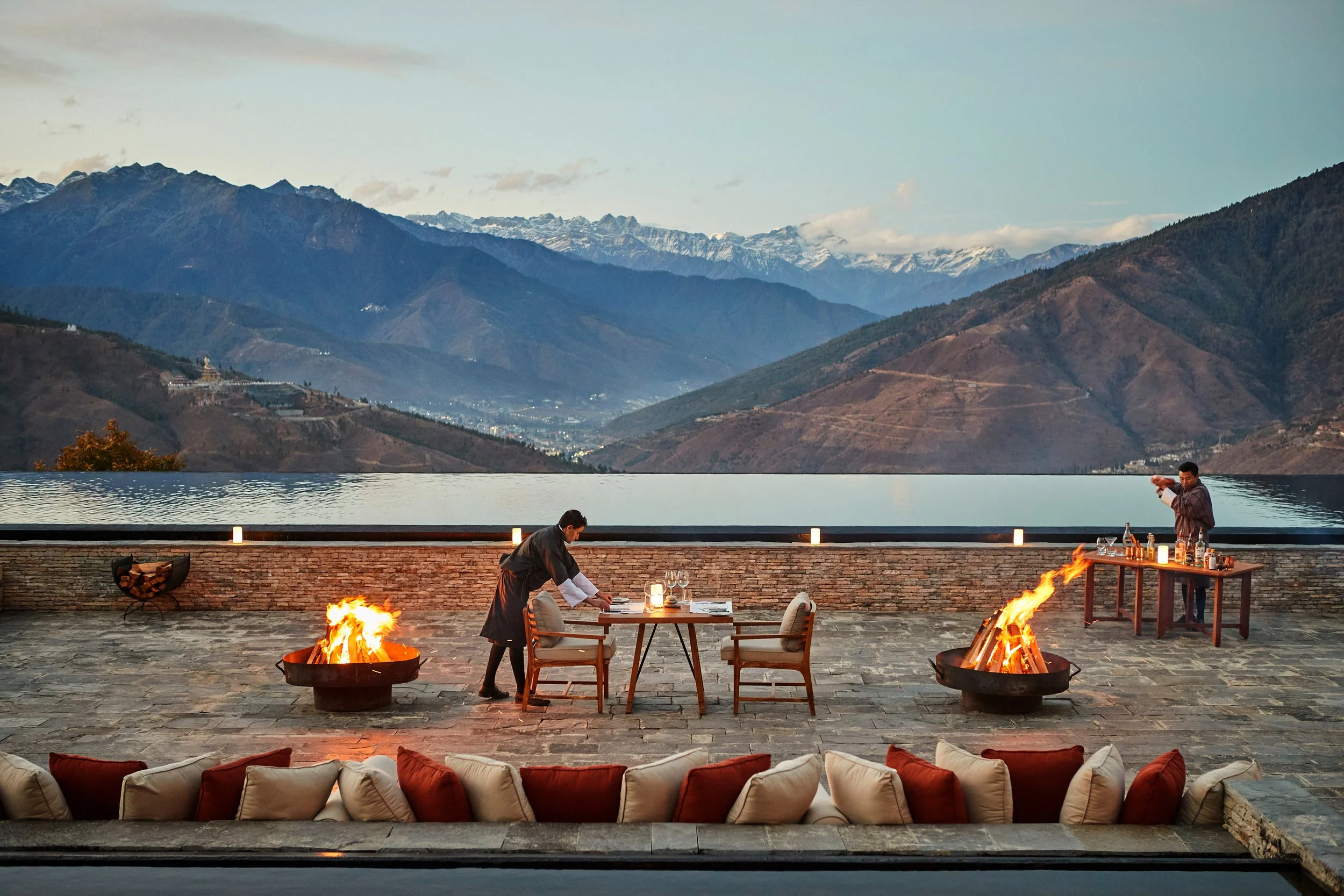 An outdoor patio with a mountain view featuring multiple fire pits, a chef preparing food at a table, and a man taking a photo, with snow-capped mountains in the background.