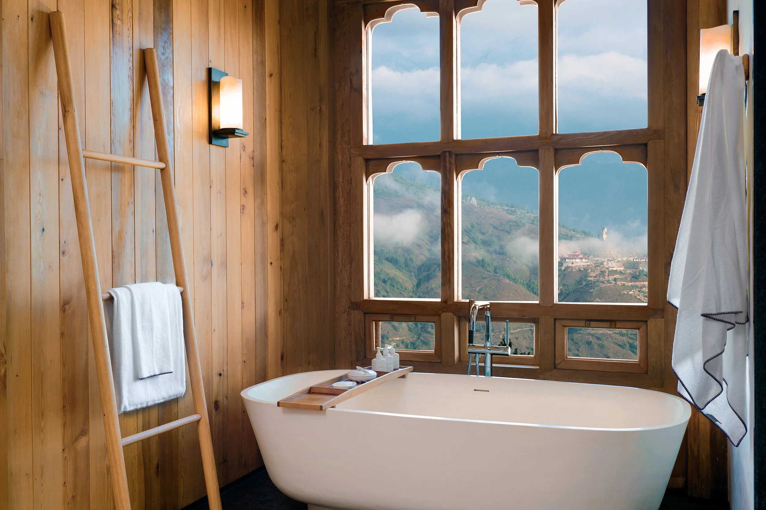 A bathroom with a freestanding bathtub placed in front of large, mountain-view windows. The walls are wooden paneling, and small shelves with toiletries are near the tub. A white towel is hanging on a wooden ladder, and another towel is hanging on a hook on the wall.