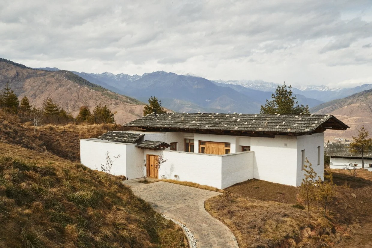 A white house with a dark roof in a mountainous area, featuring a curved stone pathway leading to the entrance, with sparse vegetation and trees around.