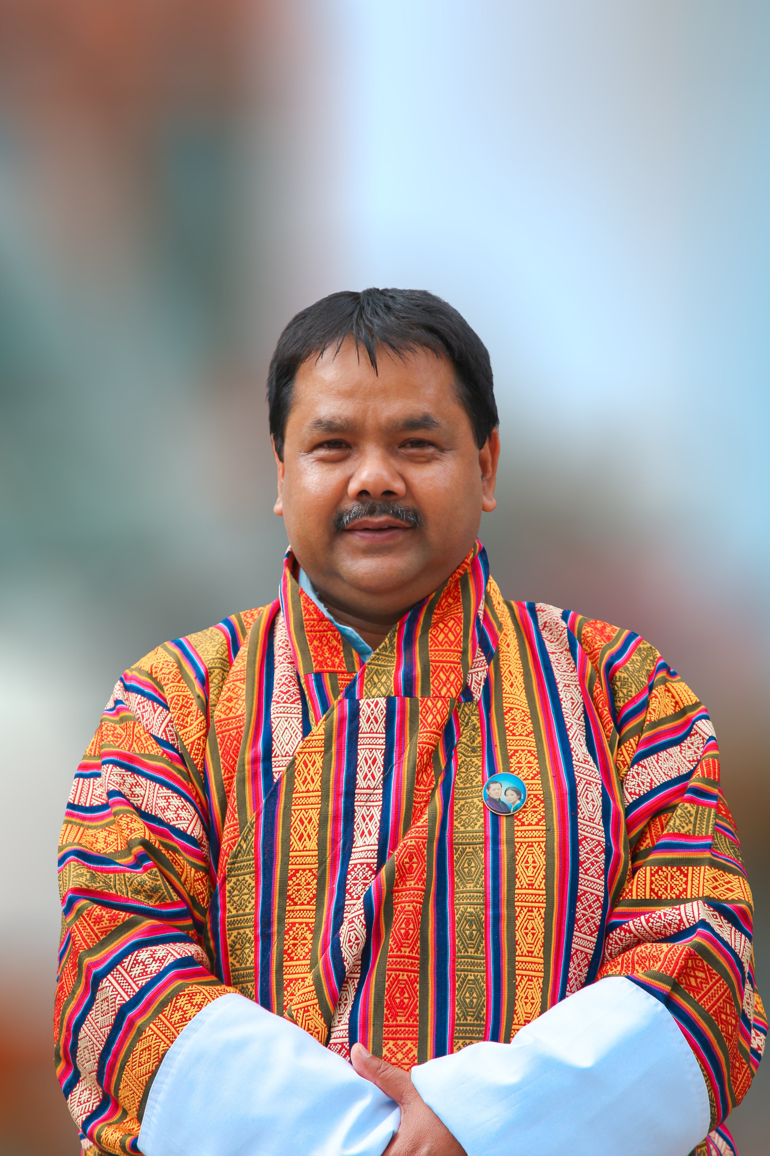 A man wearing a colorful, traditional striped textile garment, with short black hair, a mustache, and a badge pinned on his chest, standing with arms crossed.