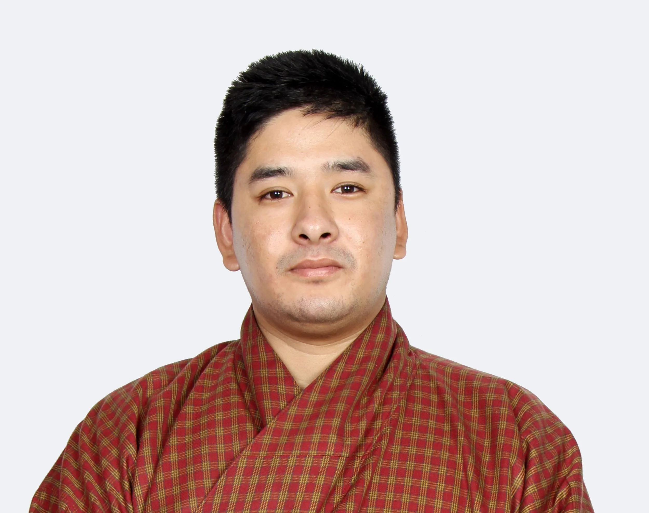 Close-up of a young man with short black hair, wearing a red and green plaid shirt, against a plain light background.