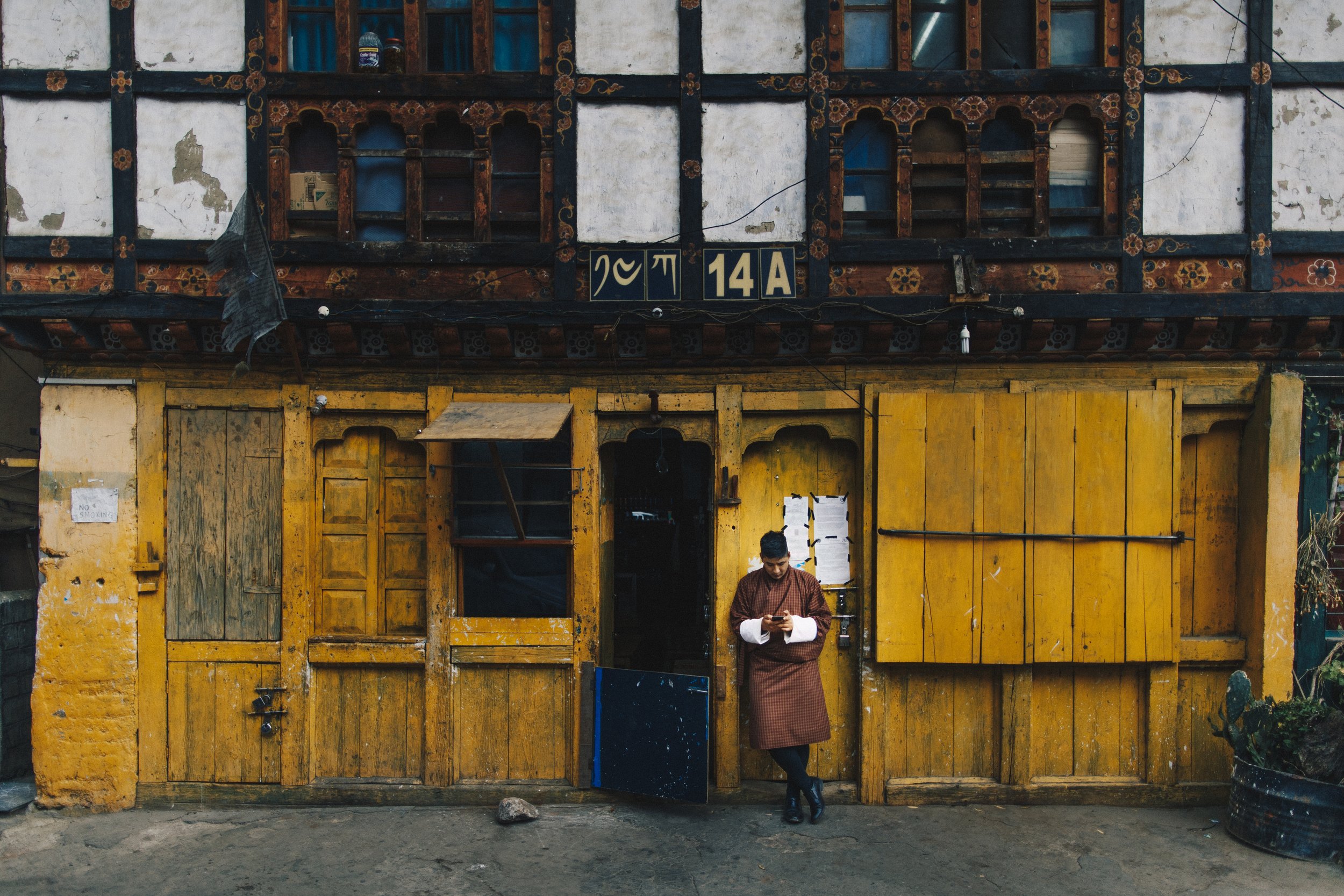A yellow wooden storefront with a person in traditional attire standing at the entrance, looking at their phone. The storefront has a black and wooden two-story facade with decorative woodwork, some windows, and signs with symbols and the address 14A.