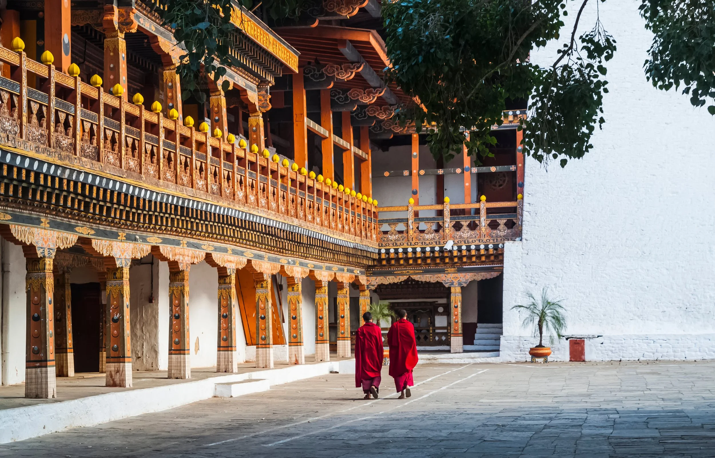 Two monks in red robes walking in front of traditional Tibetan monastery architecture with carved wooden balconies and white walls.
