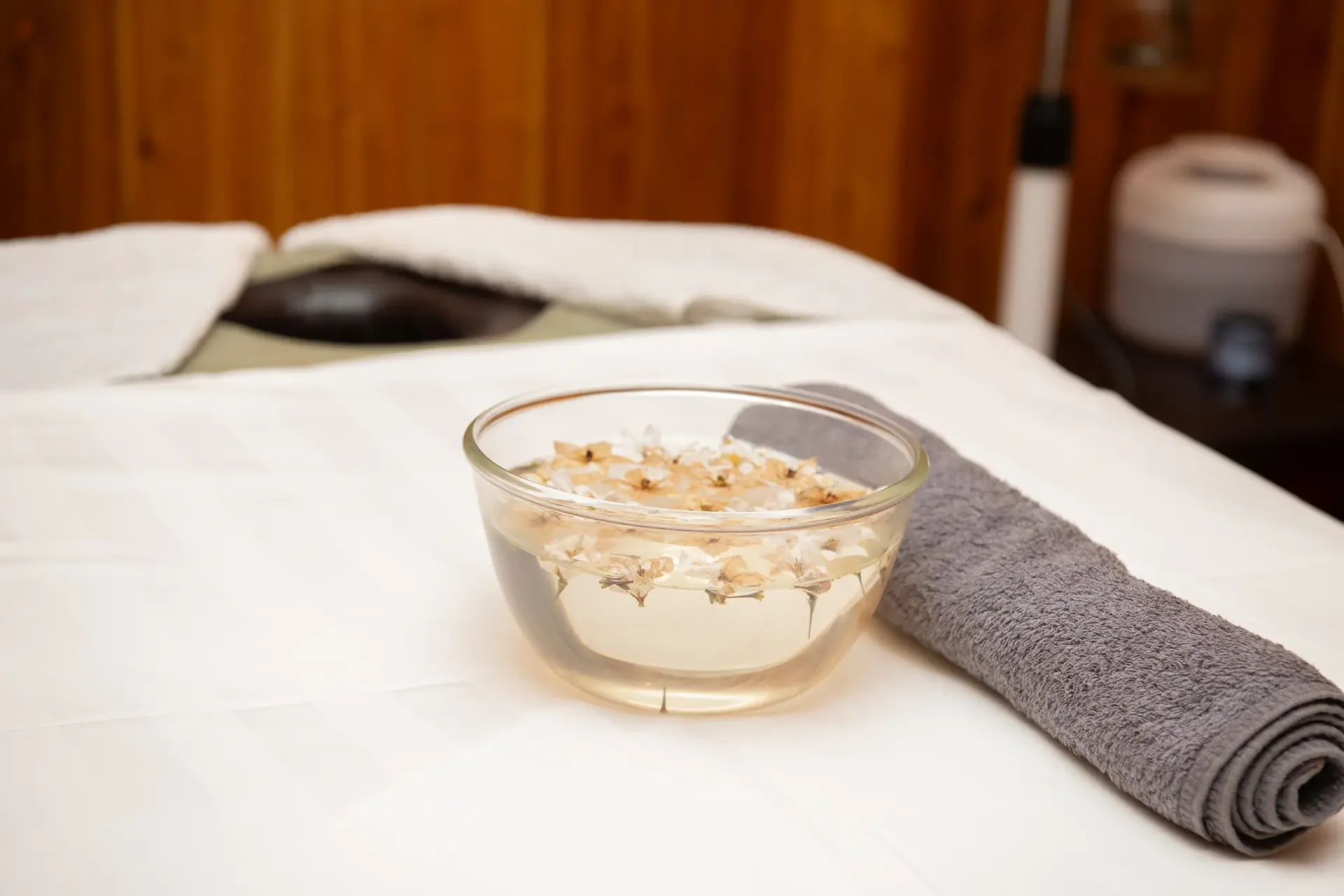 A glass bowl filled with water and floating flower petals on a table with a rolled gray towel beside it, in a room with a wooden wall in the background.