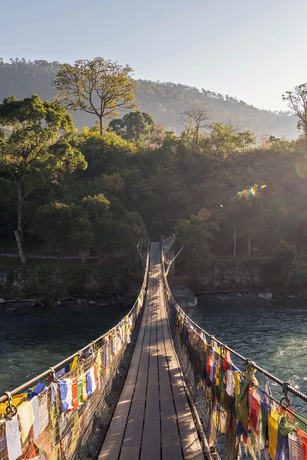 A long wooden suspension bridge with prayer flags hanging on both sides, spanning a river in a lush green forested area with mountains in the background under a clear sky.