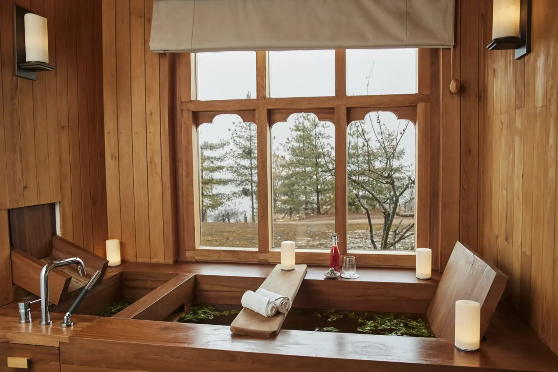 The image shows a cozy indoor space with wooden walls and a large window overlooking a tree and natural scenery. There is a small pool or hot tub with water lilies, candles, and a towel on a bench nearby. Other items include a sink, a faucet, and a kitchen or bathroom countertop.
