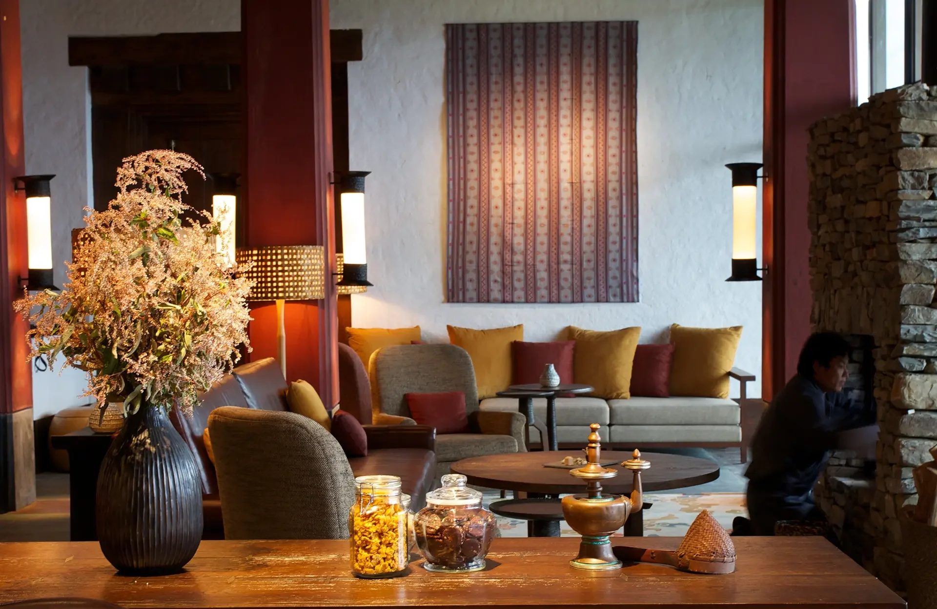 Interior of a cozy living room with a large vase of pink flowers on a wooden table, a variety of seating including a beige sofa with yellow and red cushions, warm lighting, and a stone fireplace.