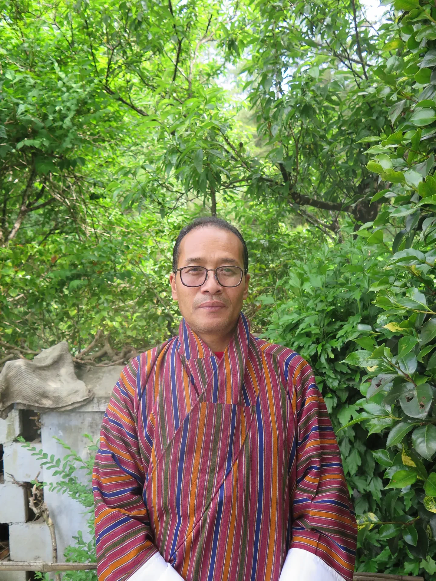 A person wearing glasses and a colorful striped traditional garment standing outdoors with lush green trees and foliage in the background.