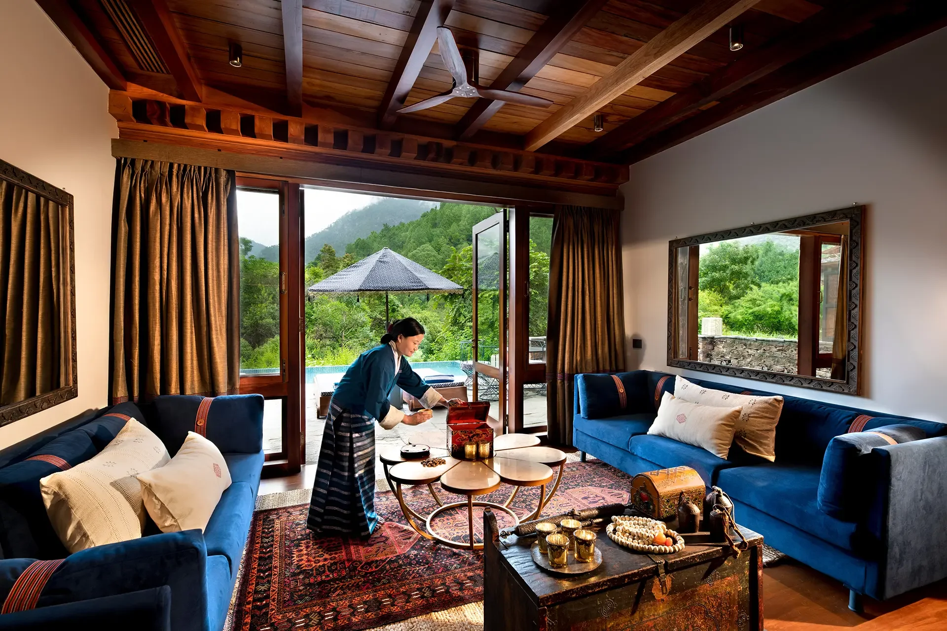 A cozy living room with large windows showing green mountains and trees outside. The room has blue sofas with pillows, a wooden coffee table with decorative items, and a patterned rug. A woman dressed in traditional attire is arranging items on the table. Curtains frame the windows, and the ceiling has wooden beams and a ceiling fan.