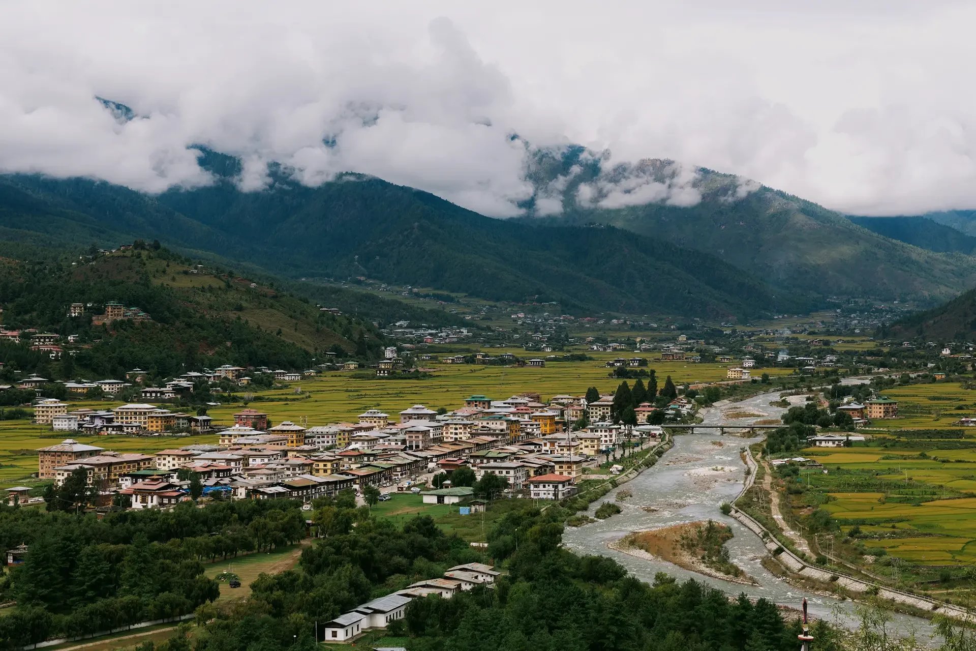 A small town in a valley with green and yellow fields, surrounded by lush green mountains, with cloudy sky overhead.