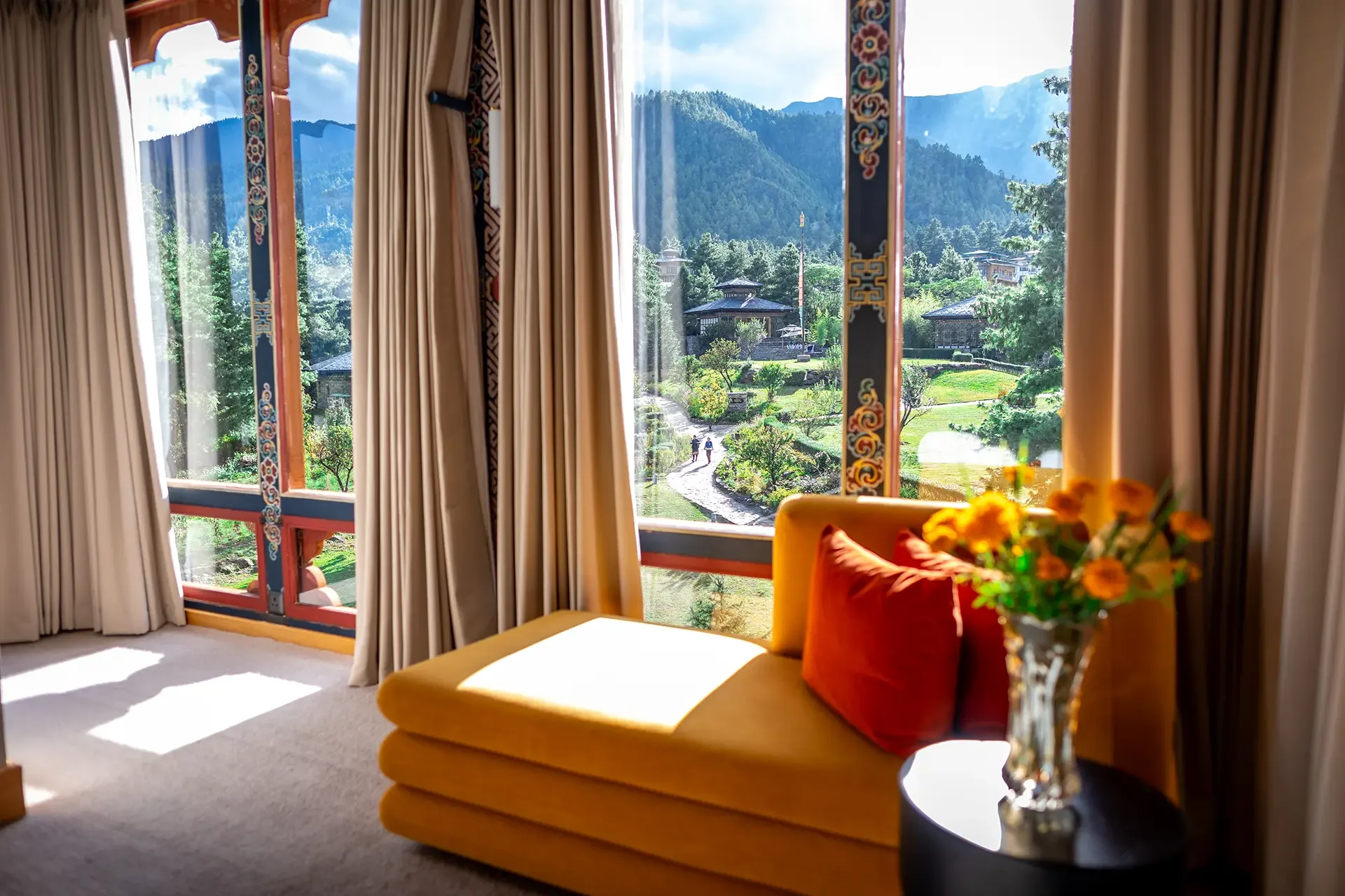 A view through a large window showing a garden with stone pathways, trees, and buildings, with mountains and a cloudy sky in the background. Inside, there are beige curtains, a yellow couch with orange and red pillows, and a vase of yellow flowers on a round black table.