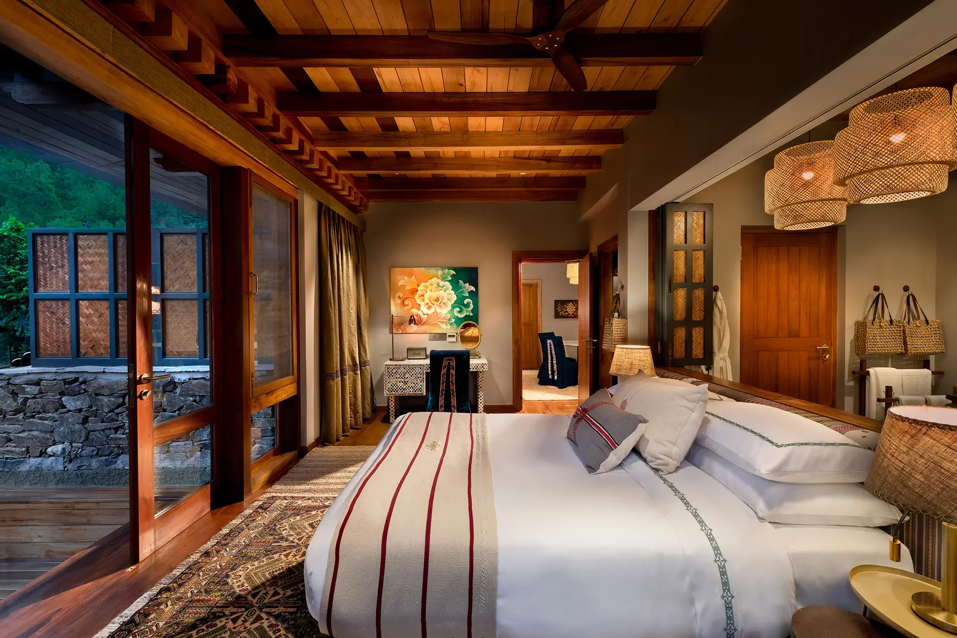 A cozy bedroom with wood accents, a bed with striped bedding, a side table with a lamp, and decorative woven lampshades hanging from the ceiling. The room has large windows showing greenery outside.