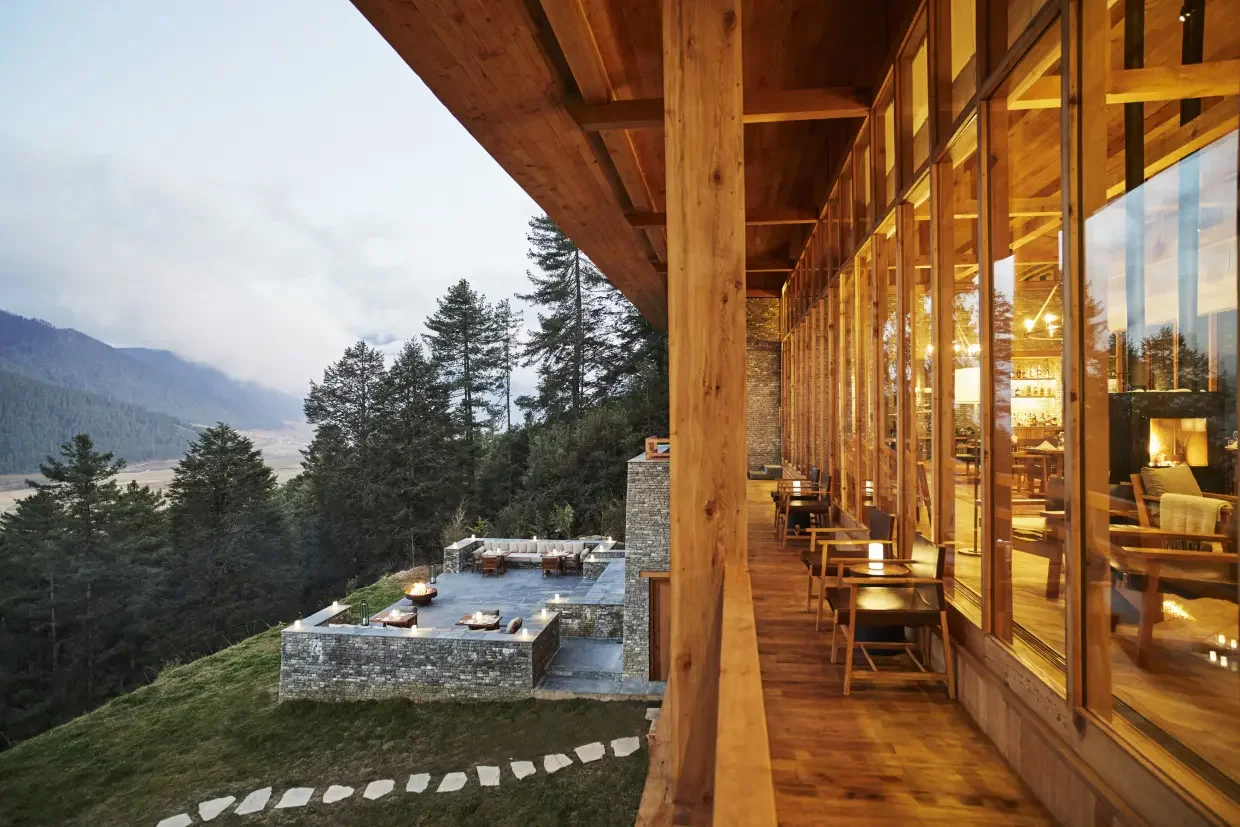 View from a glass-enclosed balcony overlooking a forested hillside and a mountain in the distance, with an outdoor patio area with seating.