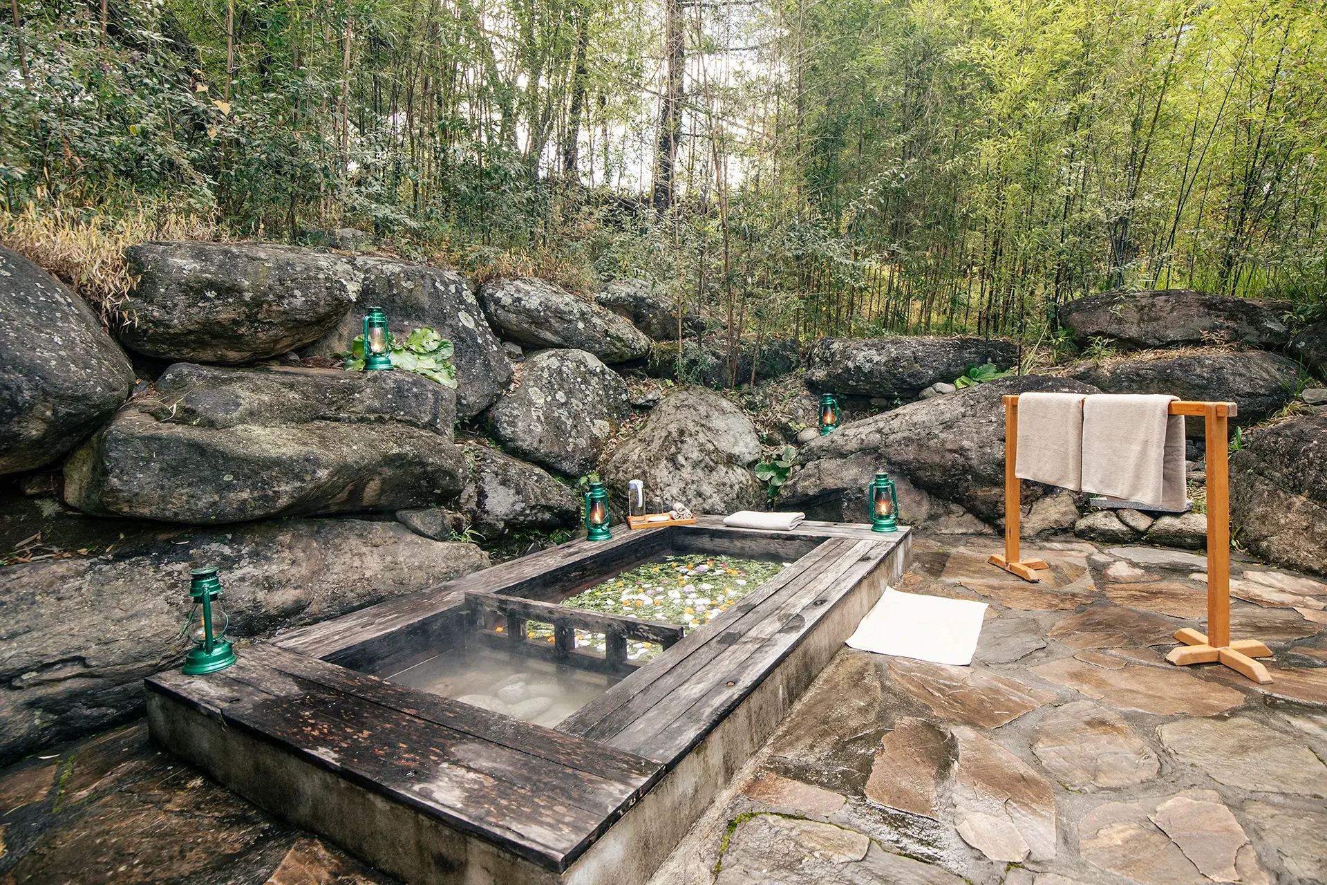 Outdoor hot spring bath with steam rising, surrounded by large rocks and lush green trees. Decorated with lanterns, towels, and floating flower petals in the water, creating a serene natural setting.