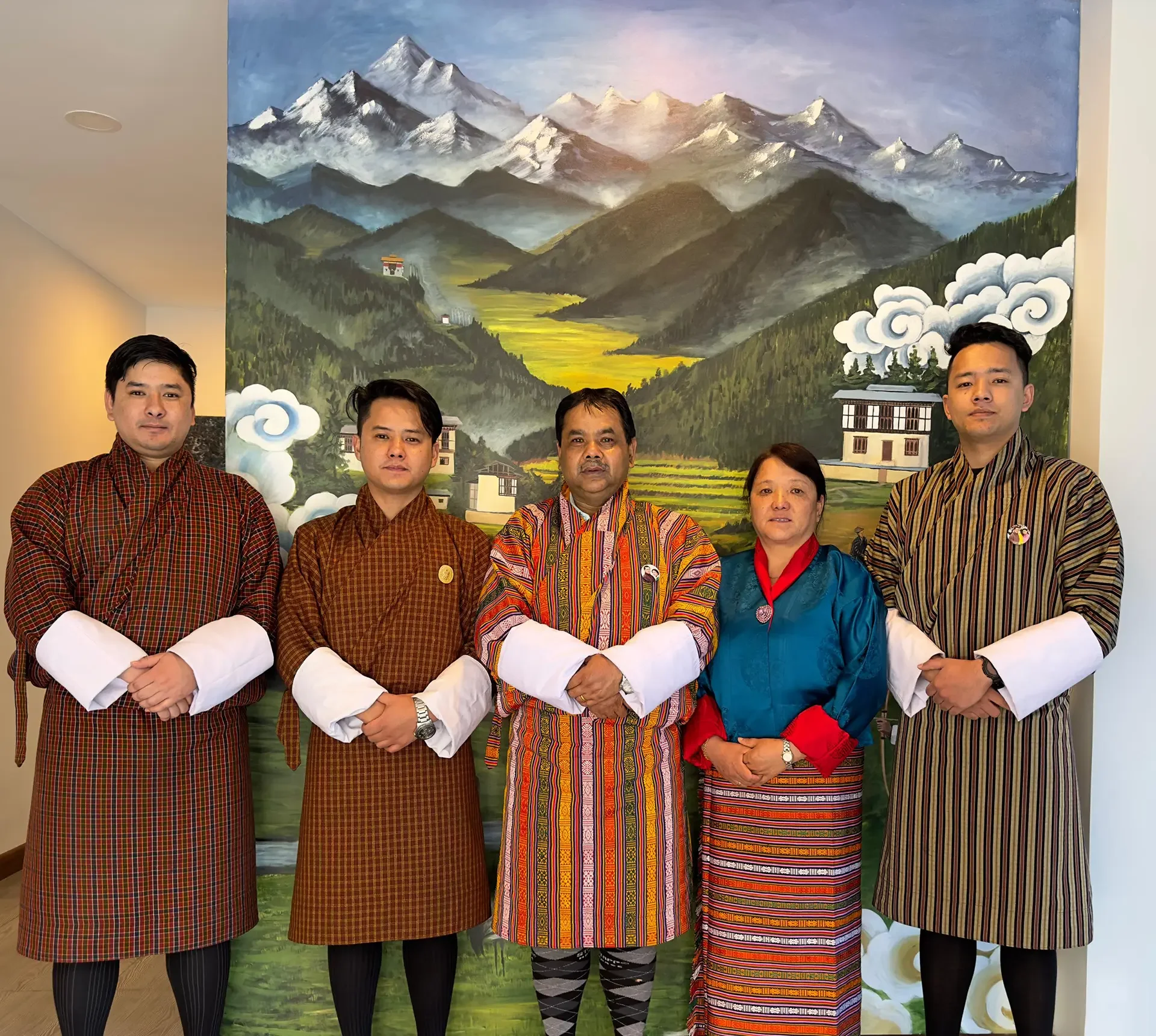 Six people standing in front of a large mountain landscape painting, wearing traditional Bhutanese clothing. The group includes both men and women, with varied expressions and postures.