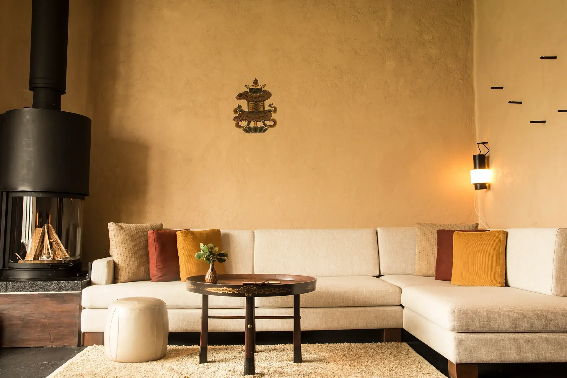 Living room with beige sectional sofa with multicolored throw pillows, a wooden coffee table with a small plant, a white ottoman, a black wood-burning stove, and warm lighting on beige textured walls.