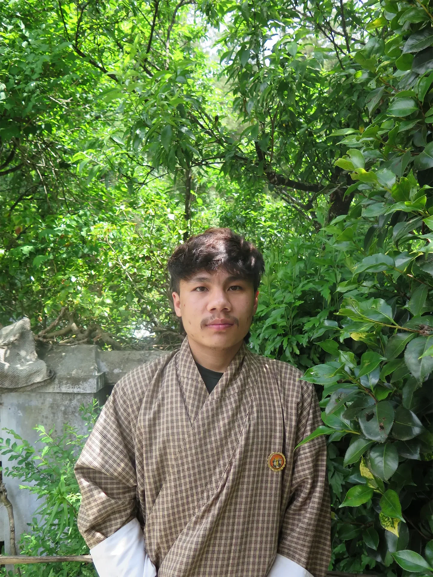 Young man with dark hair and light facial hair, wearing a brown checkered traditional robe, standing outdoors in front of dense green foliage and a concrete structure.
