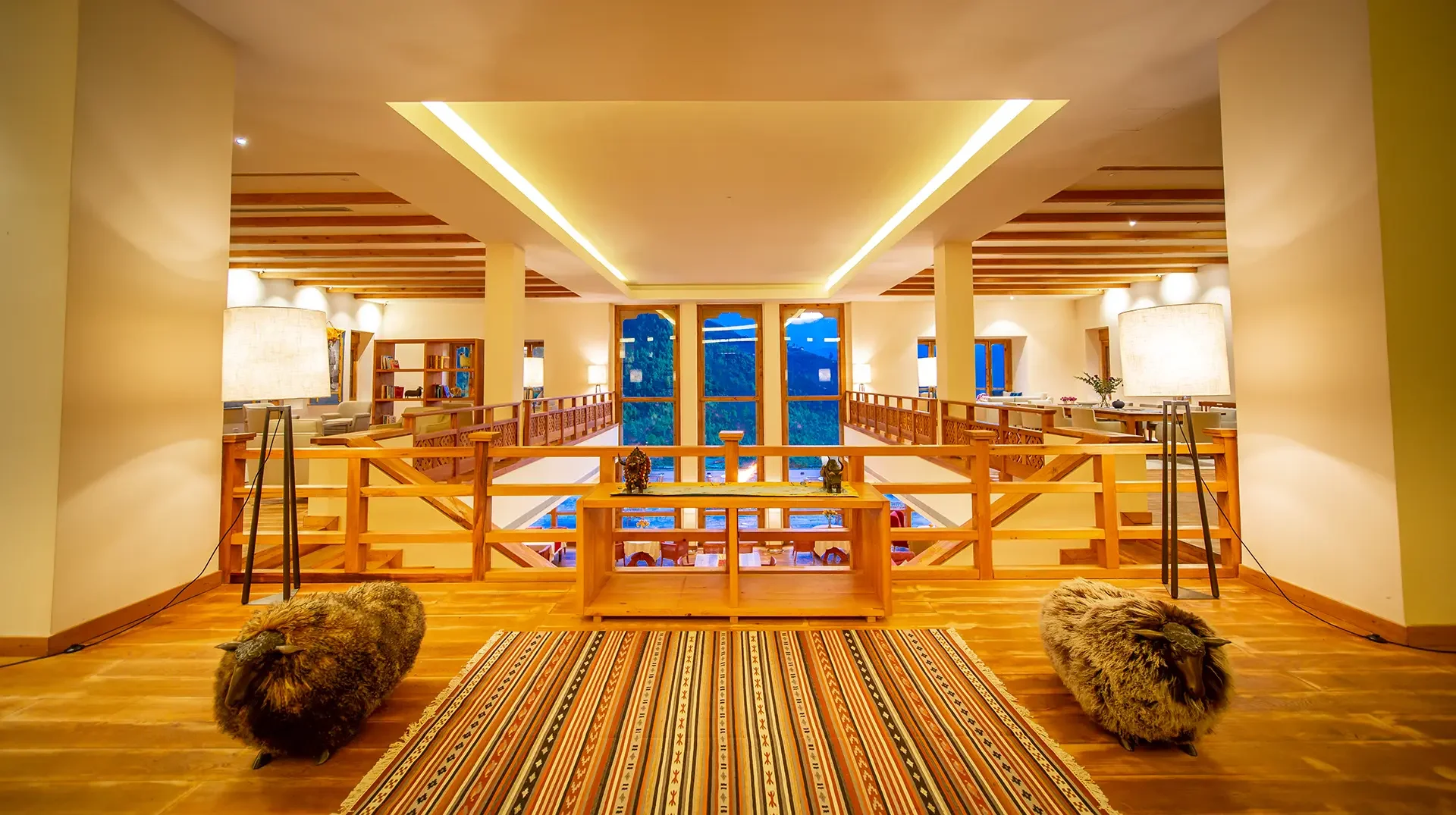 Interior of a cozy, well-lit room with wooden floors, large windows showing a mountain view, and a balcony with railings. There are two furry animal skins on the floor, a colorful striped rug, and various furniture pieces including lamps, bookshelves, and seating areas, creating a warm and inviting atmosphere.