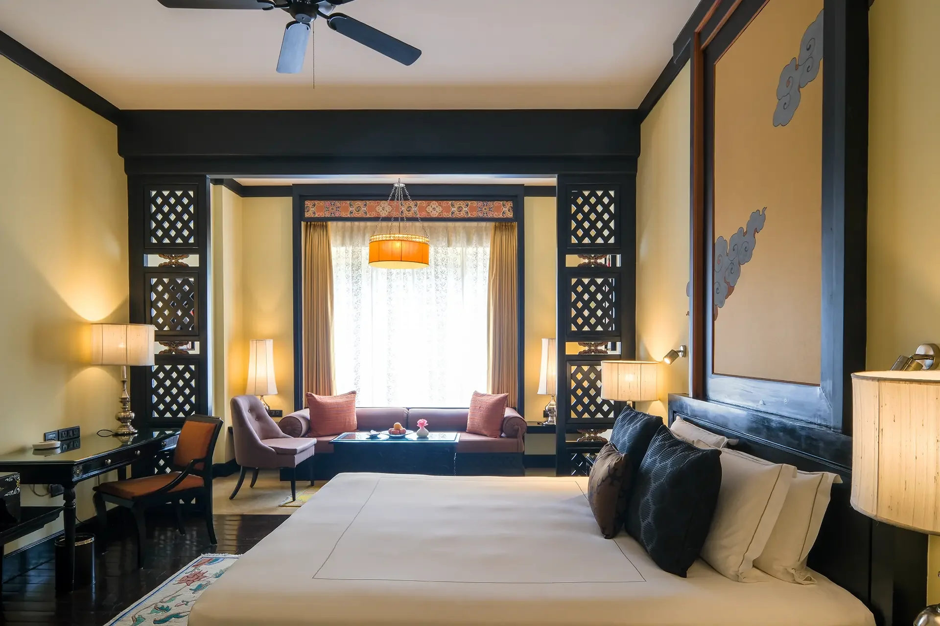 A cozy living room with yellow walls, black accents, a black ceiling fan, a chandelier, a large window with curtains, a pink sofa with cushions, lamps, and a framed artwork on the wall.