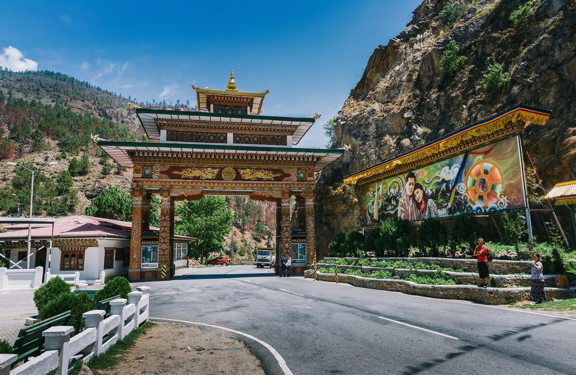 Traditional Asian-style gateway with ornate decorations and a golden pagoda roof, set against a rocky hillside with trees and a large colorful mural banner on the side.