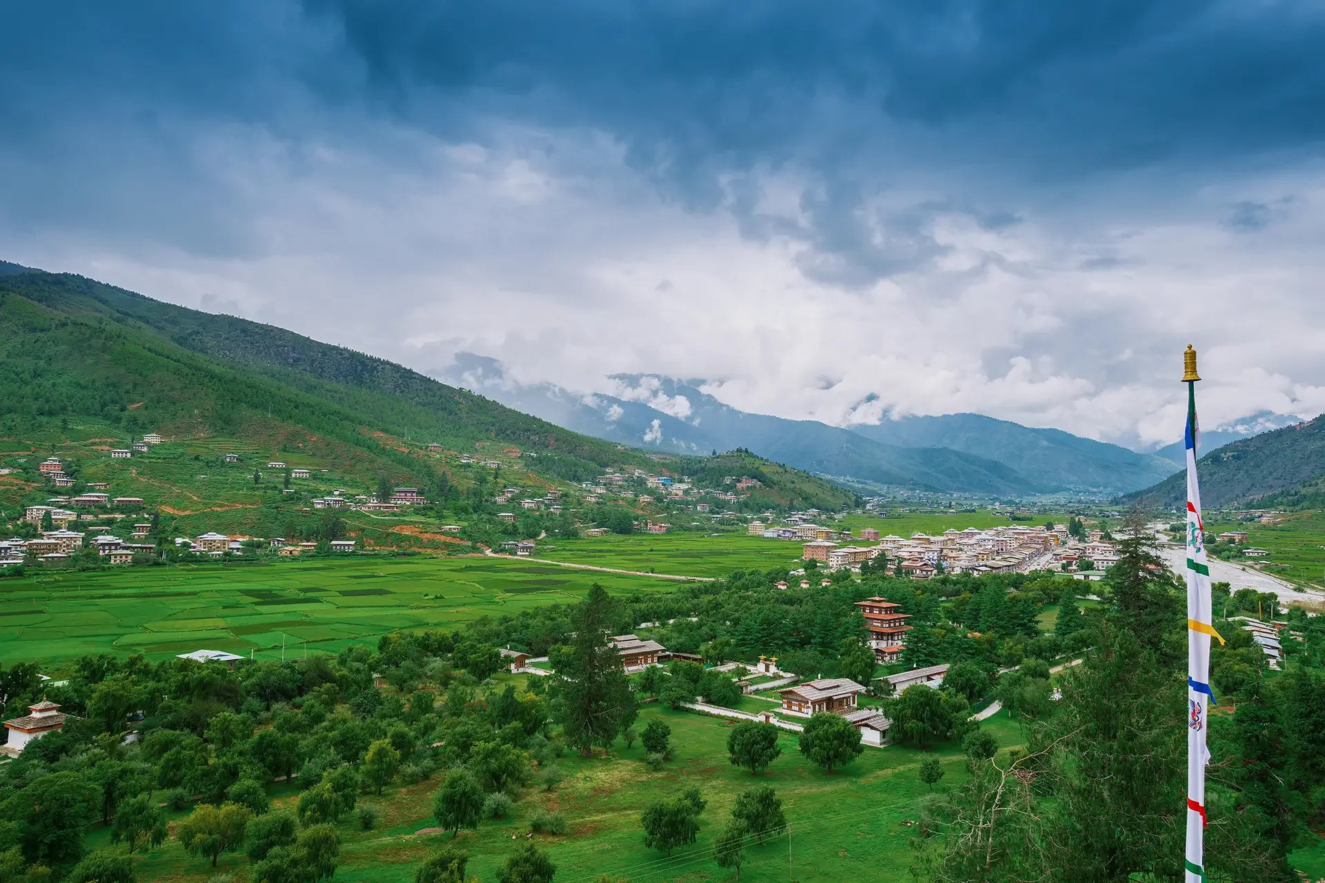 A scenic landscape of mountains with lush green terraced fields in the foreground and a village with houses and buildings scattered along the foothills. A flagpole with a colorful flag stands on the right side, and the sky is cloudy with patches of blue.
