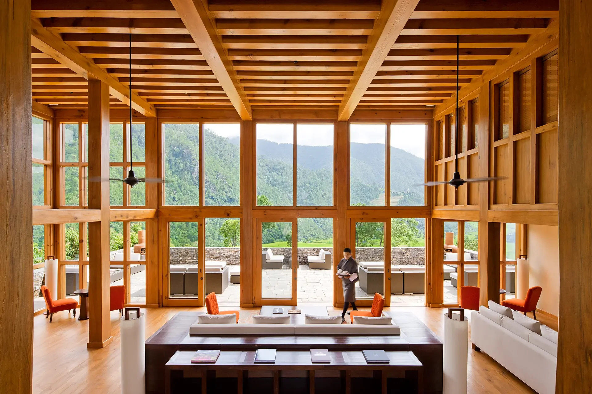 Interior view of a modern wooden lodge with large windows overlooking a lush green mountain landscape. The space features wooden beams and ceiling, with a seating area including white sofas and orange chairs. A staff member is walking in the background near the outdoor patio.