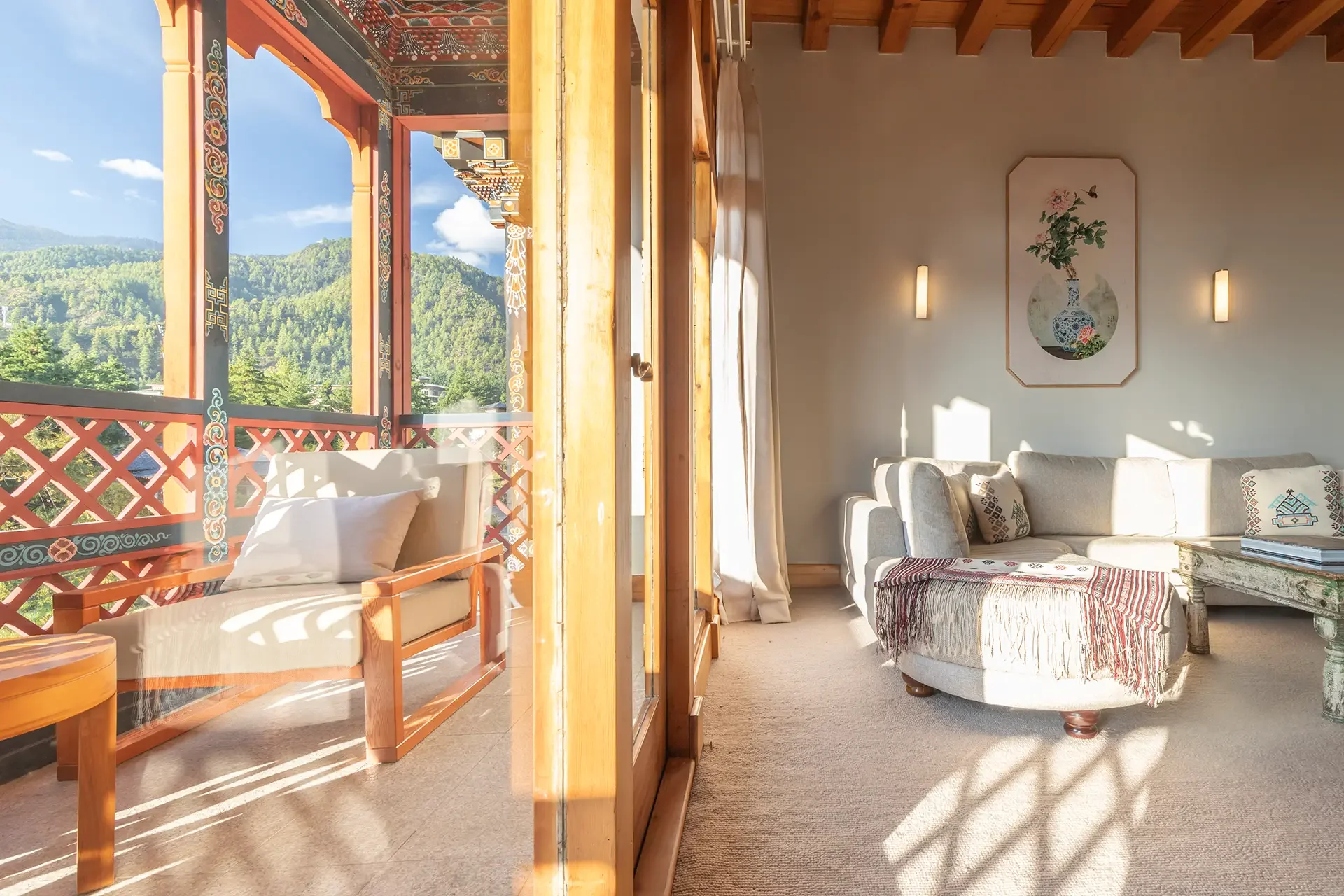 Bright living room with large sliding glass doors leading to a balcony with mountain view, furnished with a beige sofa with decorative pillows, artwork on the wall, and sunlight streaming in.