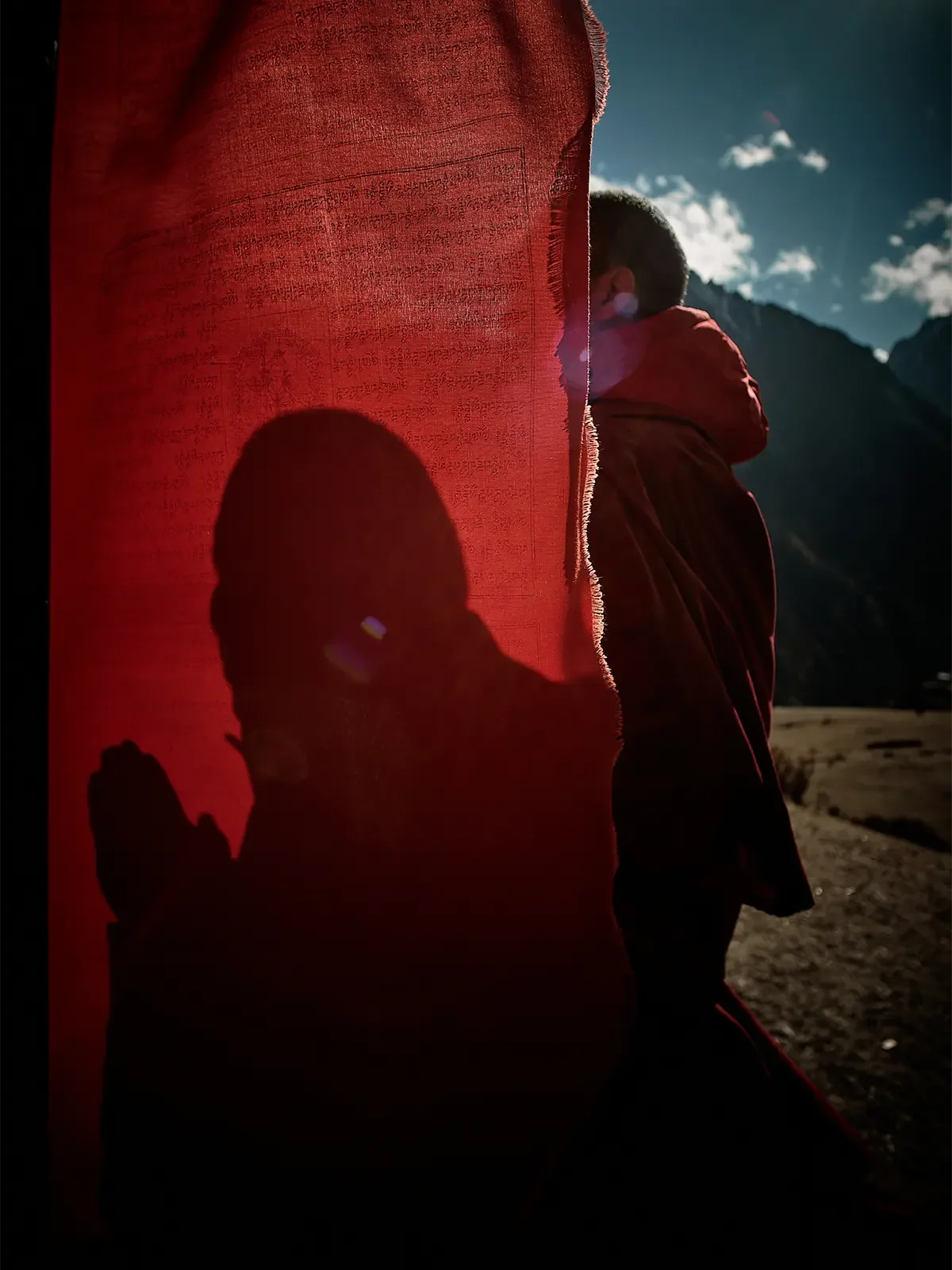 A person covered with a red cloth standing outdoors with mountains in the background, with a strong shadow of another person on the red cloth, under a partly cloudy sky.