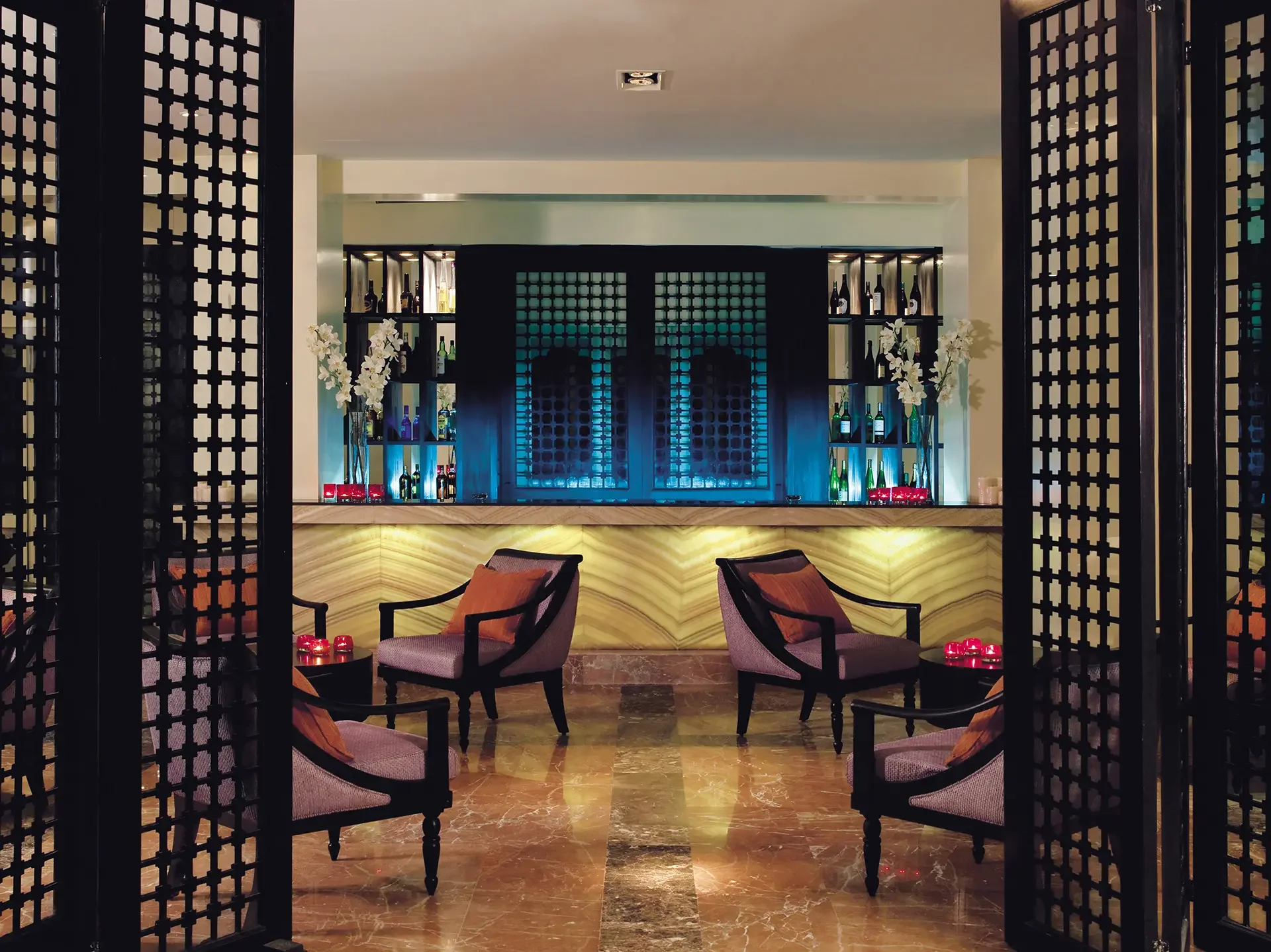 A luxurious hotel lobby or lounge area with a bar in the background. The space features a marble floor, stylish chairs with cushions, decorative planters with white flowers, and a dark wooden partition with a geometric pattern framing the view.