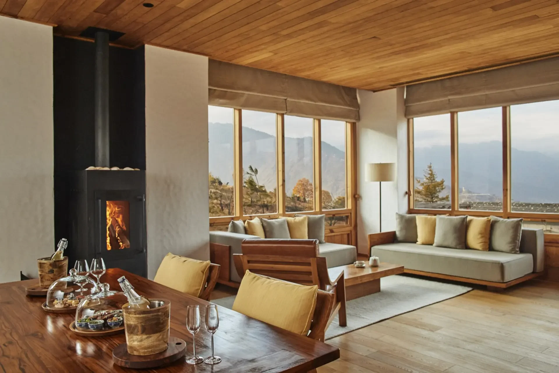 Interior view of a living room with large windows overlooking mountains and trees, with a cozy fireplace on the left, sofas with pillows, a wooden floor, and a lamp.