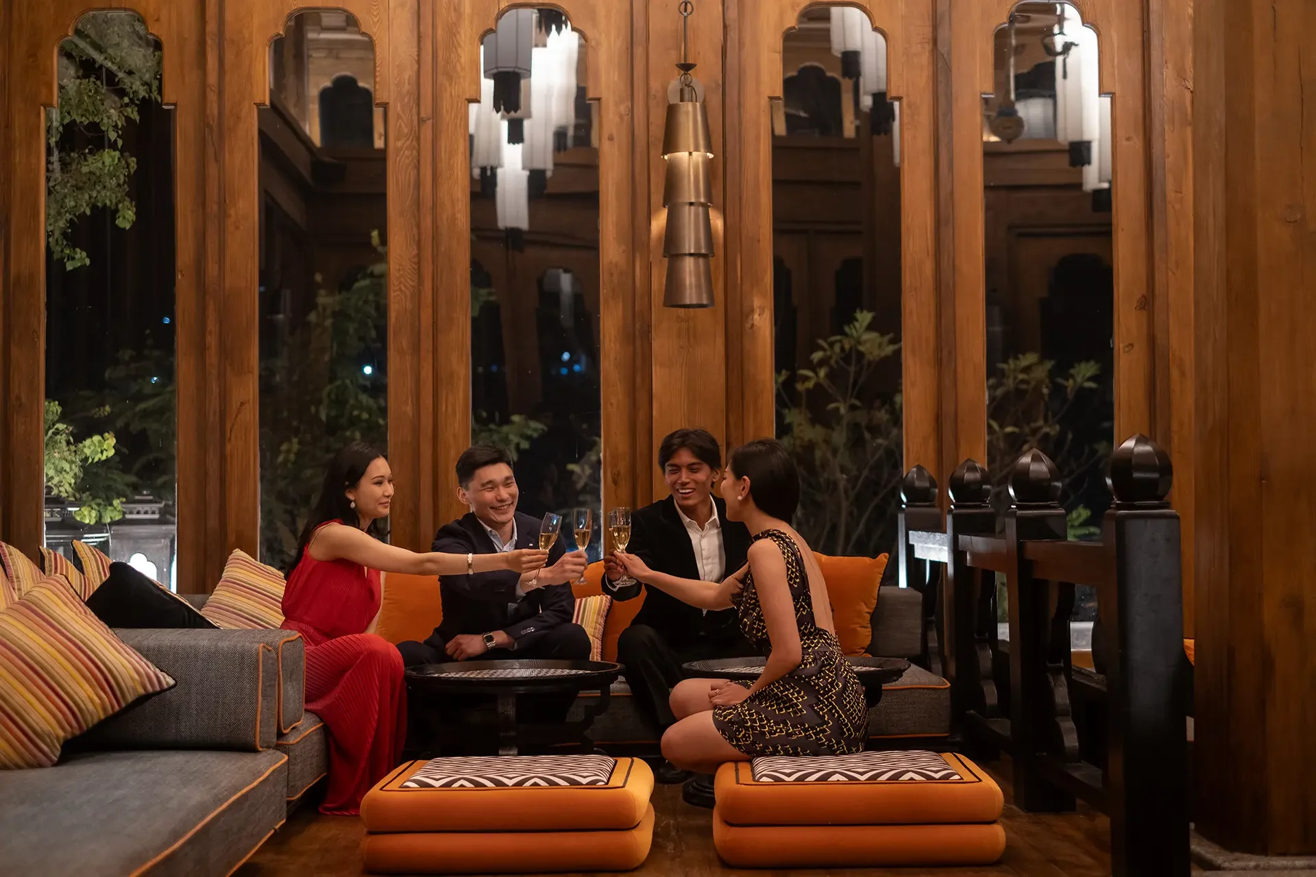 A group of five people sitting on cushions in an elegant wooden room at night, raising glasses of champagne in a toast, smiling and enjoying each other's company.
