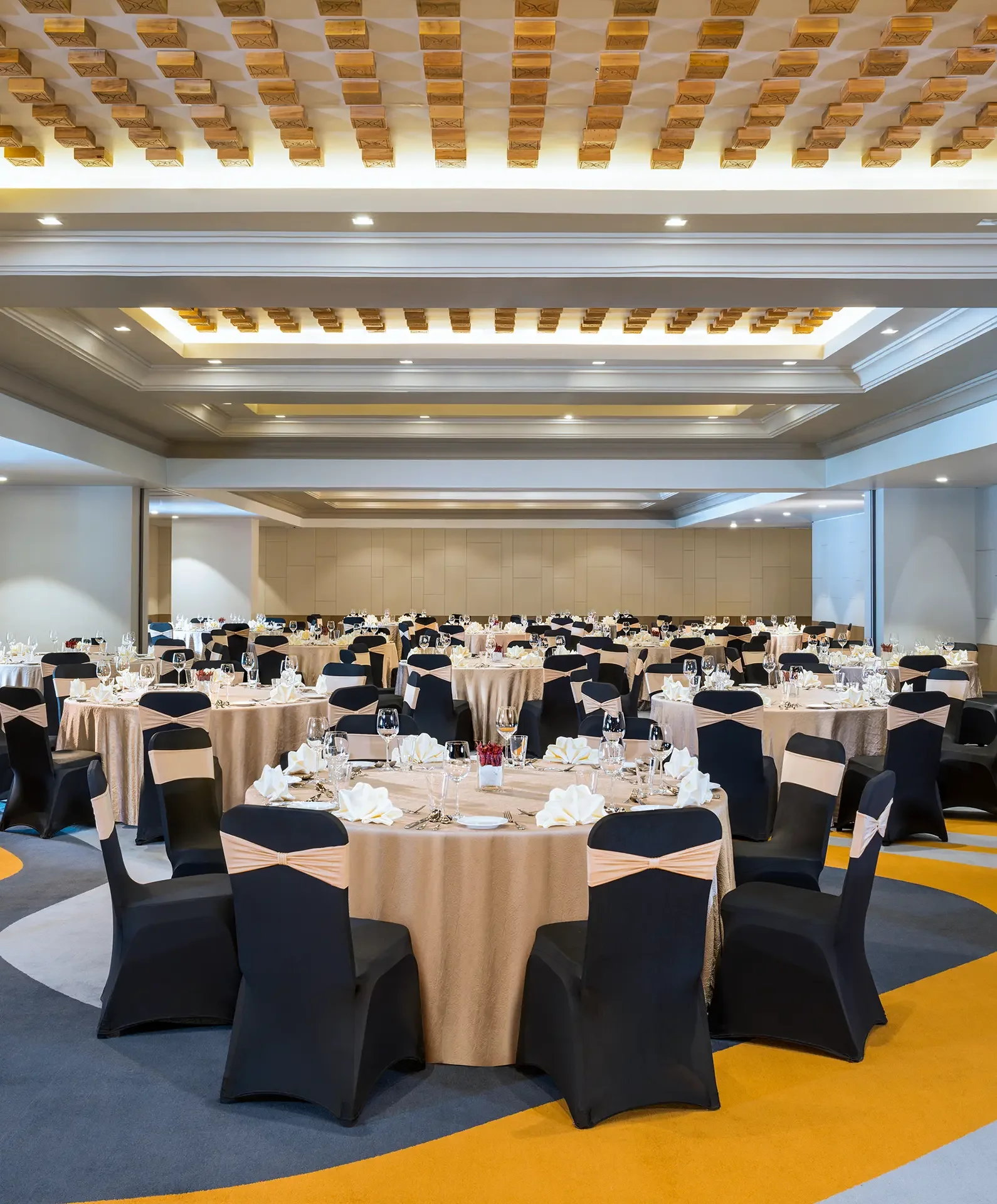 A banquet hall with round tables covered in beige tablecloths, set with glassware, napkins, and centerpieces. Black chairs with beige sashes surround each table. The ceiling features decorative wooden beams and recessed lighting, and the floor has a colorful geometric pattern.