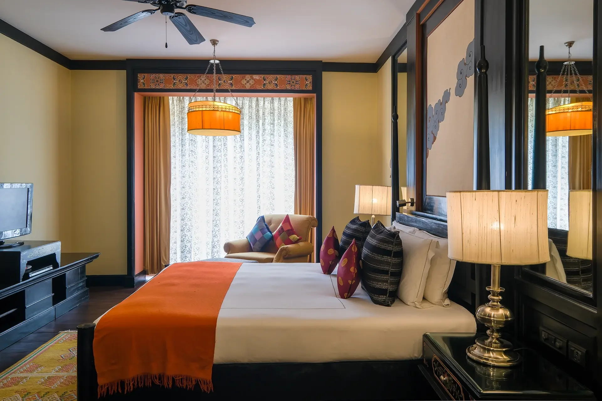 A bedroom with a large bed covered with white sheets, orange blanket, and orange bed runner. Decorative pillows are arranged on the bed. There is a nightstand with a brass lamp on one side, and a small TV and dresser on the other. The room has yellow walls, dark wood furniture, and a window with lace curtains and orange drapes. A ceiling fan is installed on the ceiling, and a lounge chair with multicolored cushions is near the window.